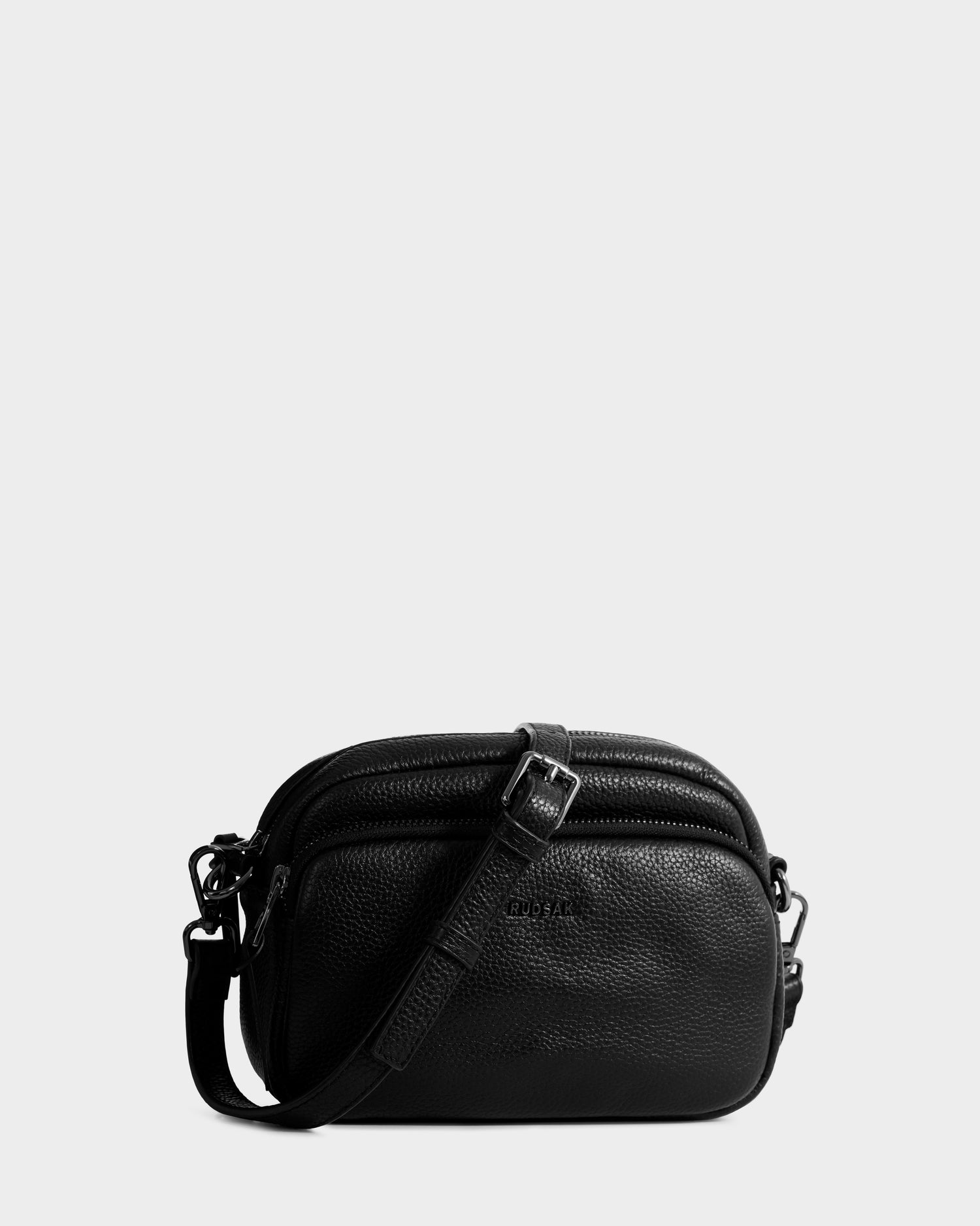 The ZOYA - L325015 Black by Rudsak is a small women’s crossbody bag in pebbled leather, featuring a textured finish, dual zippered compartments, and an adjustable strap, shown on a plain light gray background.