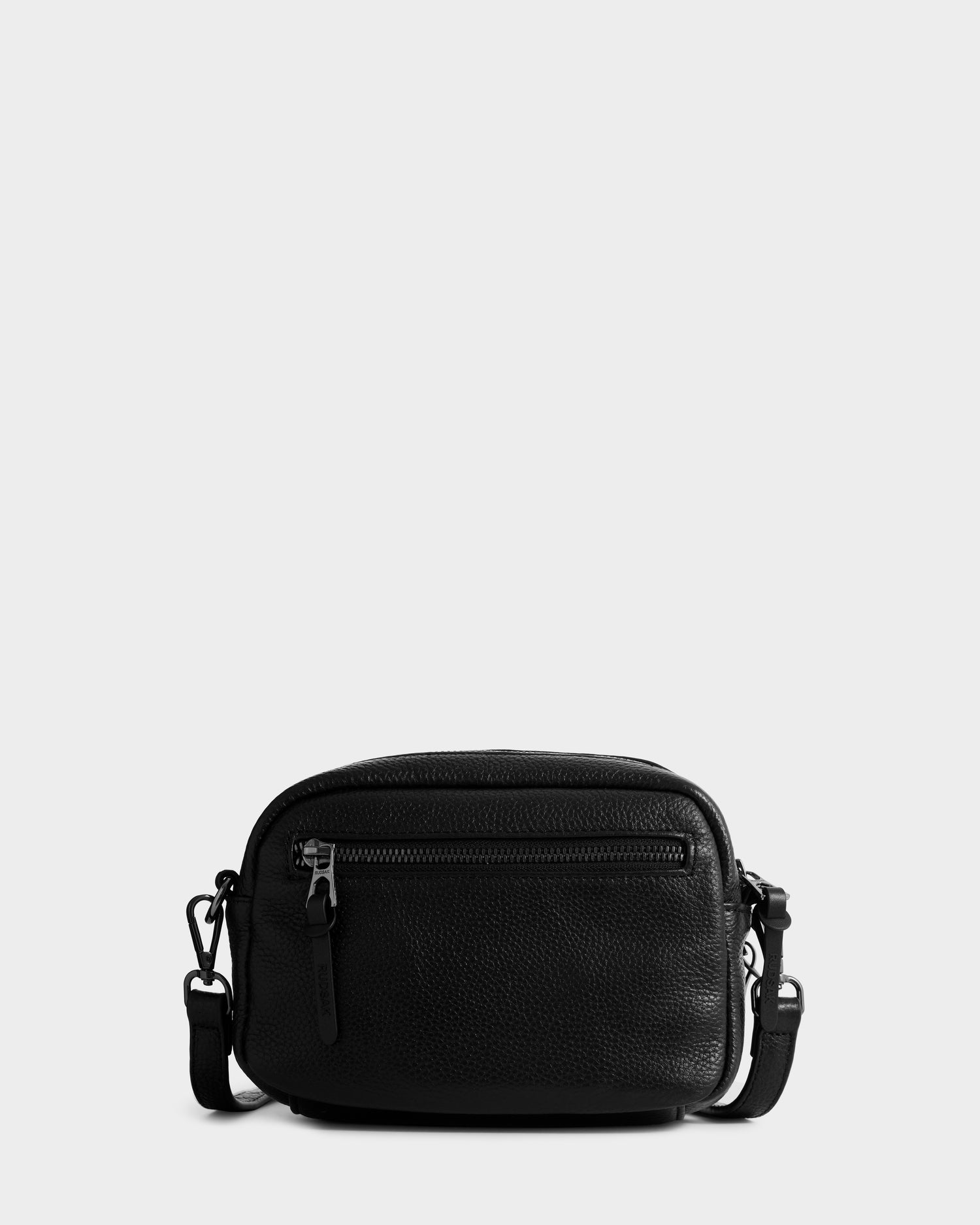 The Rudsak ZOYA - L325015 Black is a small women’s crossbody bag in black pebbled leather with a front zipper pocket, adjustable strap, and minimalist design, shown against a plain light gray background.