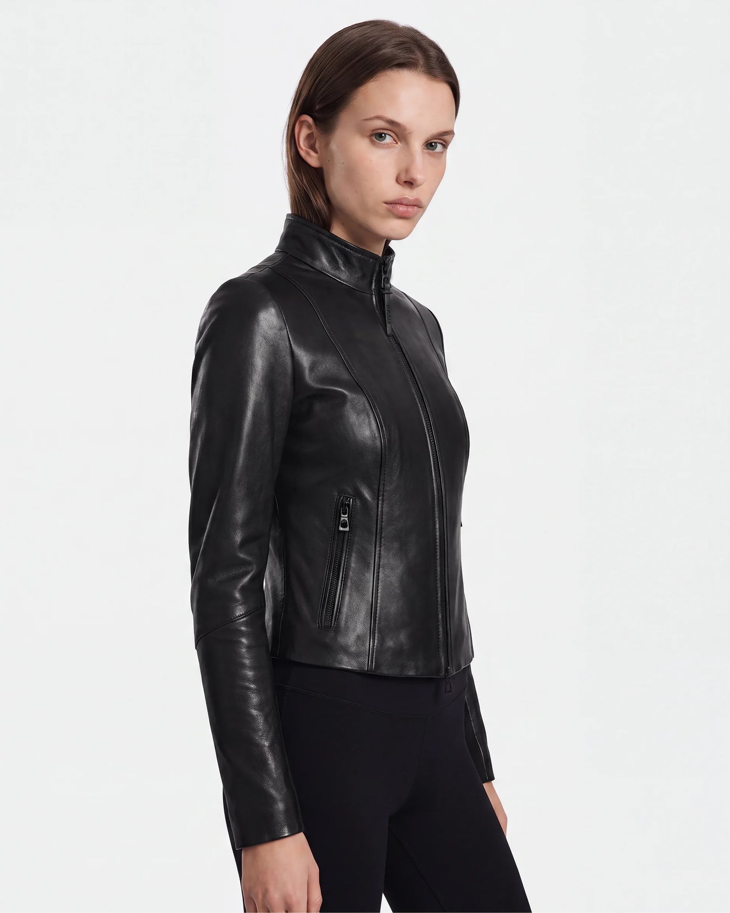 A woman with straight brown hair wears the ABIGAIL - 8124560 Black leather racer jacket by Rudsak and black pants, standing against a plain white background and looking slightly to the side with a neutral expression.