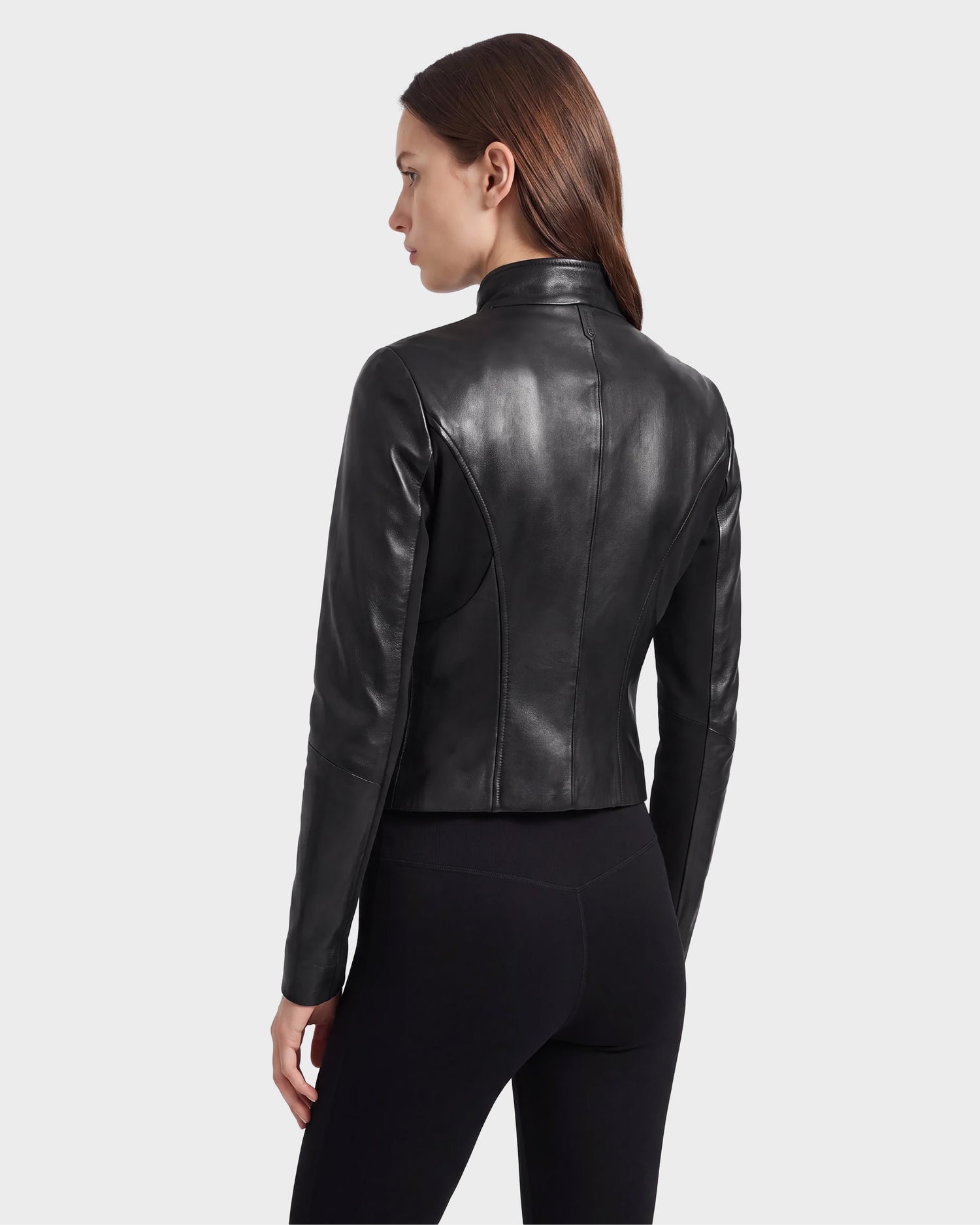 A woman with straight brown hair is shown from behind, wearing the ABIGAIL - 8124560 Black women’s leather racer jacket by Rudsak paired with black pants, standing against a plain light gray background.