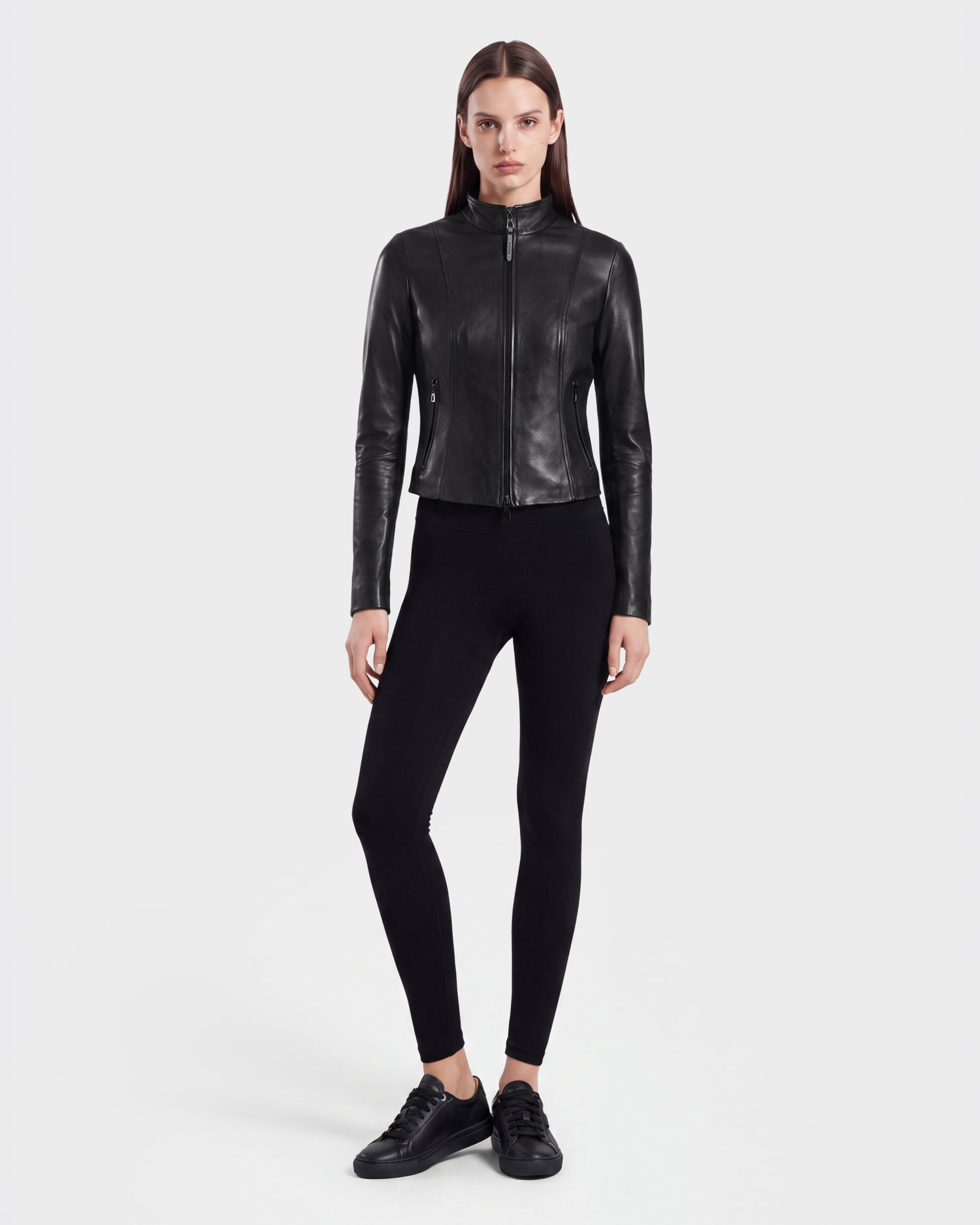 A woman with straight brown hair and a neutral expression stands against a plain background, dressed in the ABIGAIL - 8124560 Black leather racer jacket by Rudsak. She pairs the fitted black women's jacket with black leggings and black sneakers.