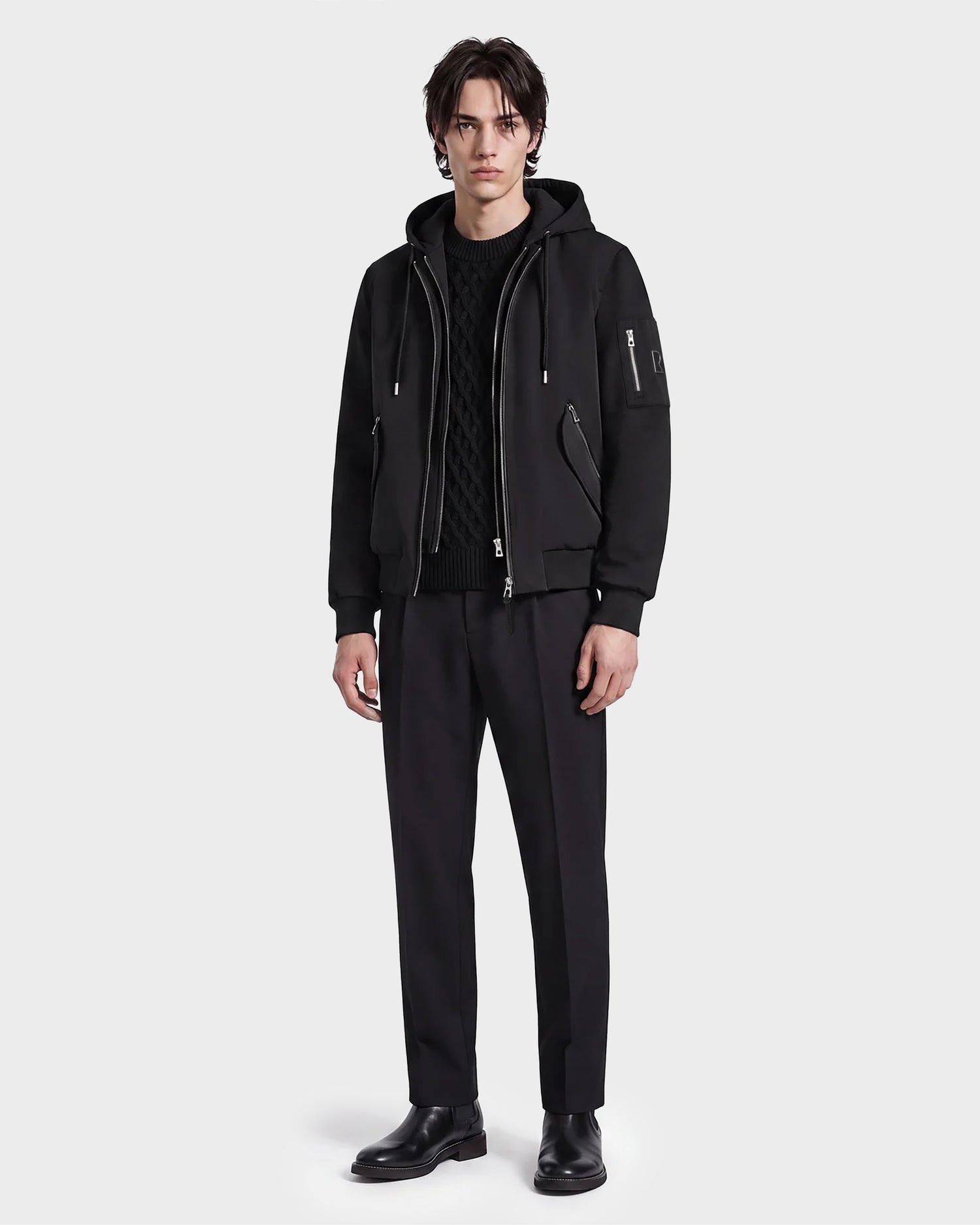 A man with dark hair and a neutral expression stands against a white background, wearing the Rudsak ANDRE - 6124020 Black men's lightweight bomber jacket with a removable hooded bib. He pairs it with a black sweater, black trousers, and black boots, perfectly capturing a modern style.