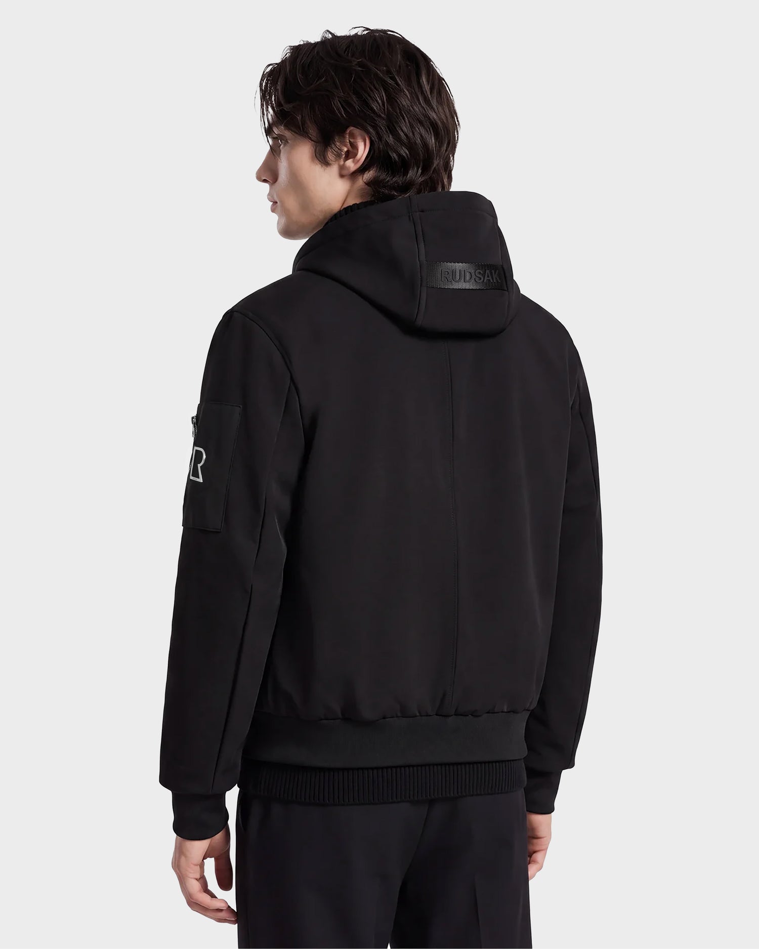 A person with medium-length dark hair stands with their back to the camera against a plain light background, wearing the Rudsak ANDRE - 6124020 Black modern bomber jacket with a removable hooded bib and black pants.