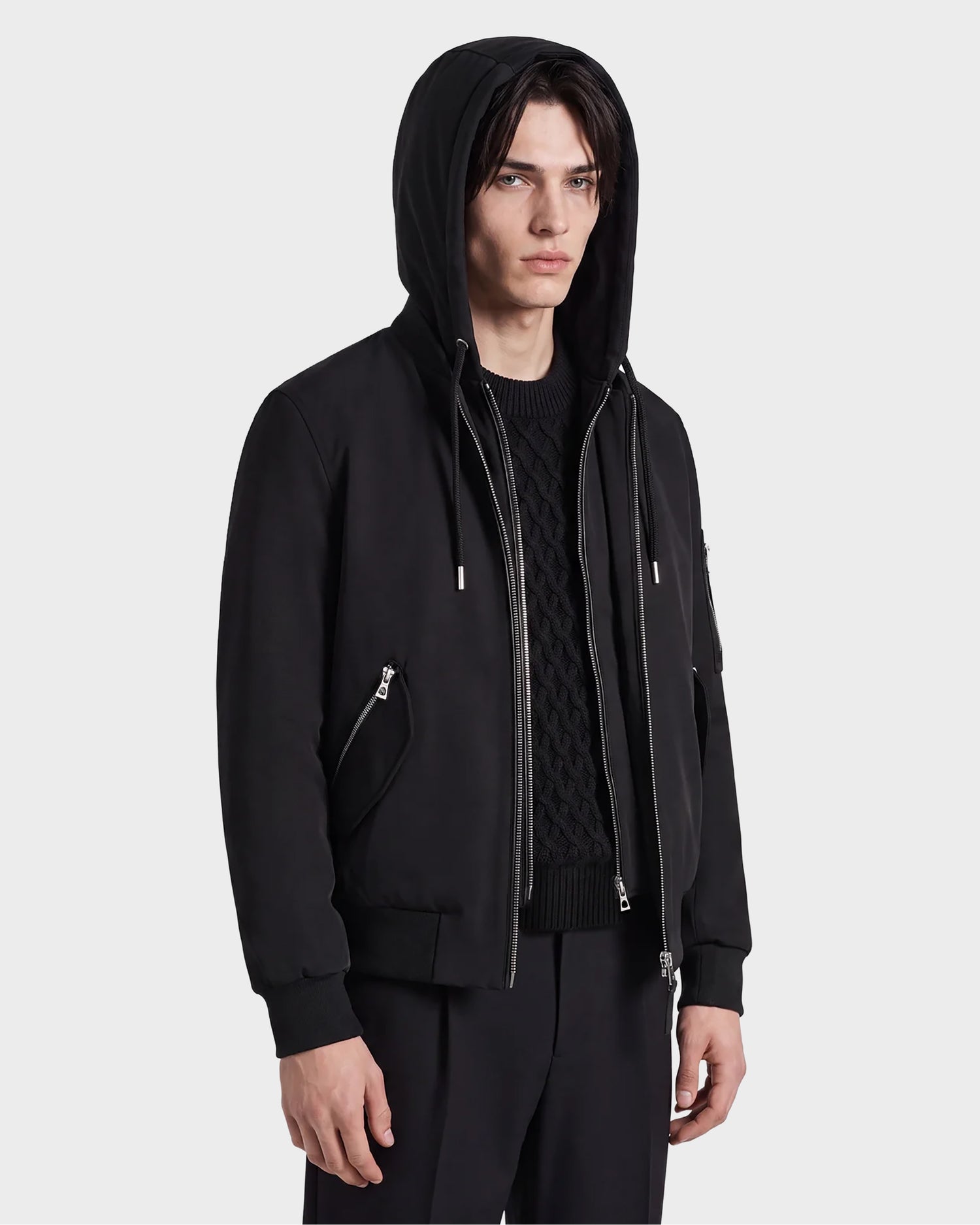A person wearing the Rudsak ANDRE - 6124020 Black men's lightweight bomber jacket with a removable hooded bib over a black textured sweater and black pants stands facing slightly to the right with a neutral expression, showcasing a modern style.