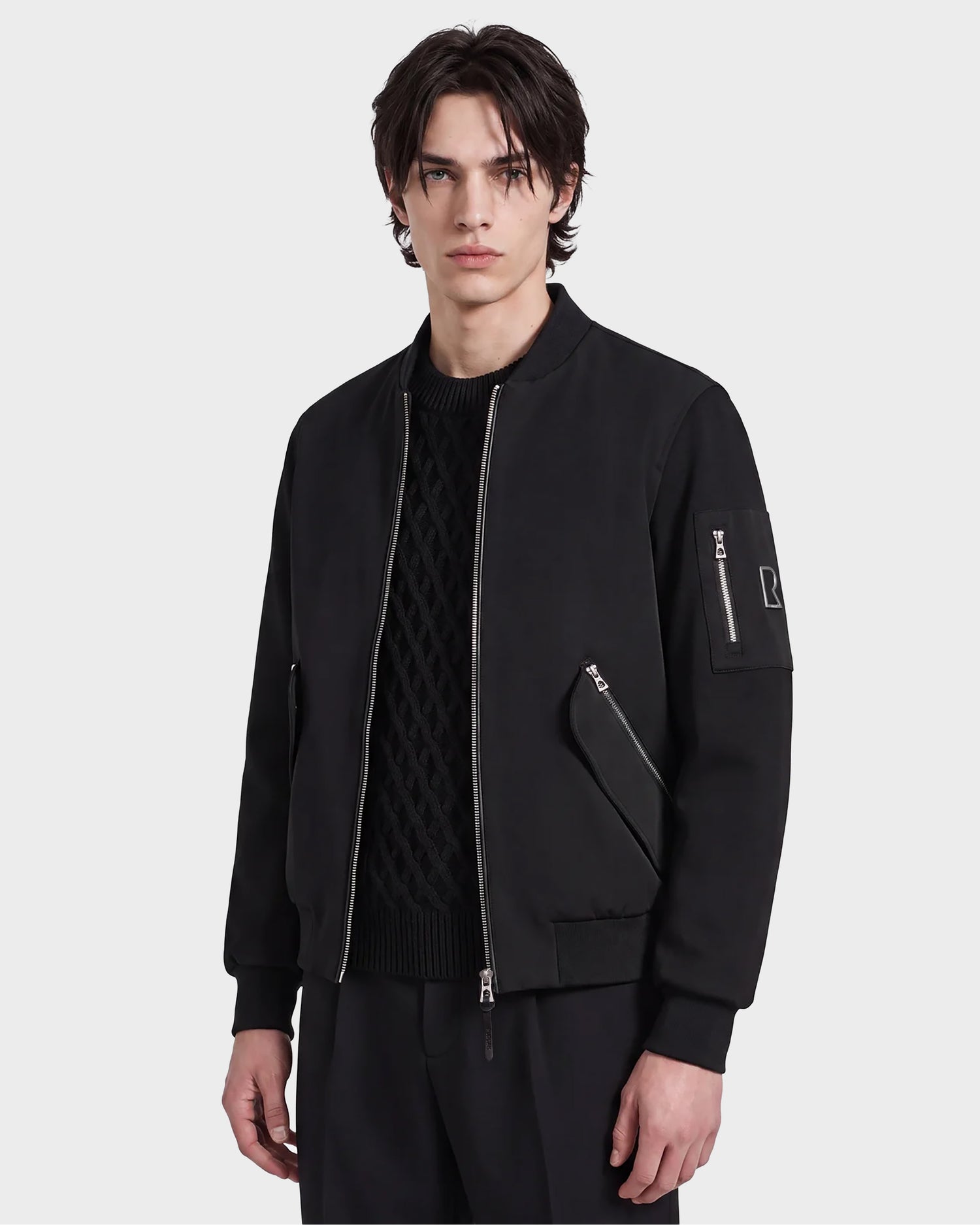 A young man with medium-length dark hair wears the Rudsak ANDRE - 6124020 Black modern bomber jacket with a removable hooded bib layered over a textured black sweater, standing against a plain white background.