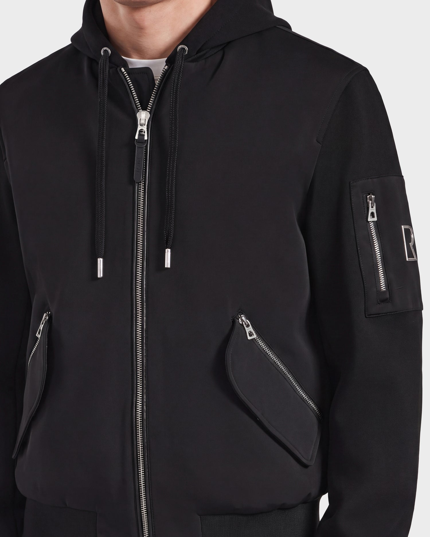 A person wearing the ANDRE - 6124020 Black men's lightweight bomber jacket by Rudsak, featuring a modern style with silver front and sleeve zippers, angled zippered pockets, and a removable hooded bib. Only the torso is visible.