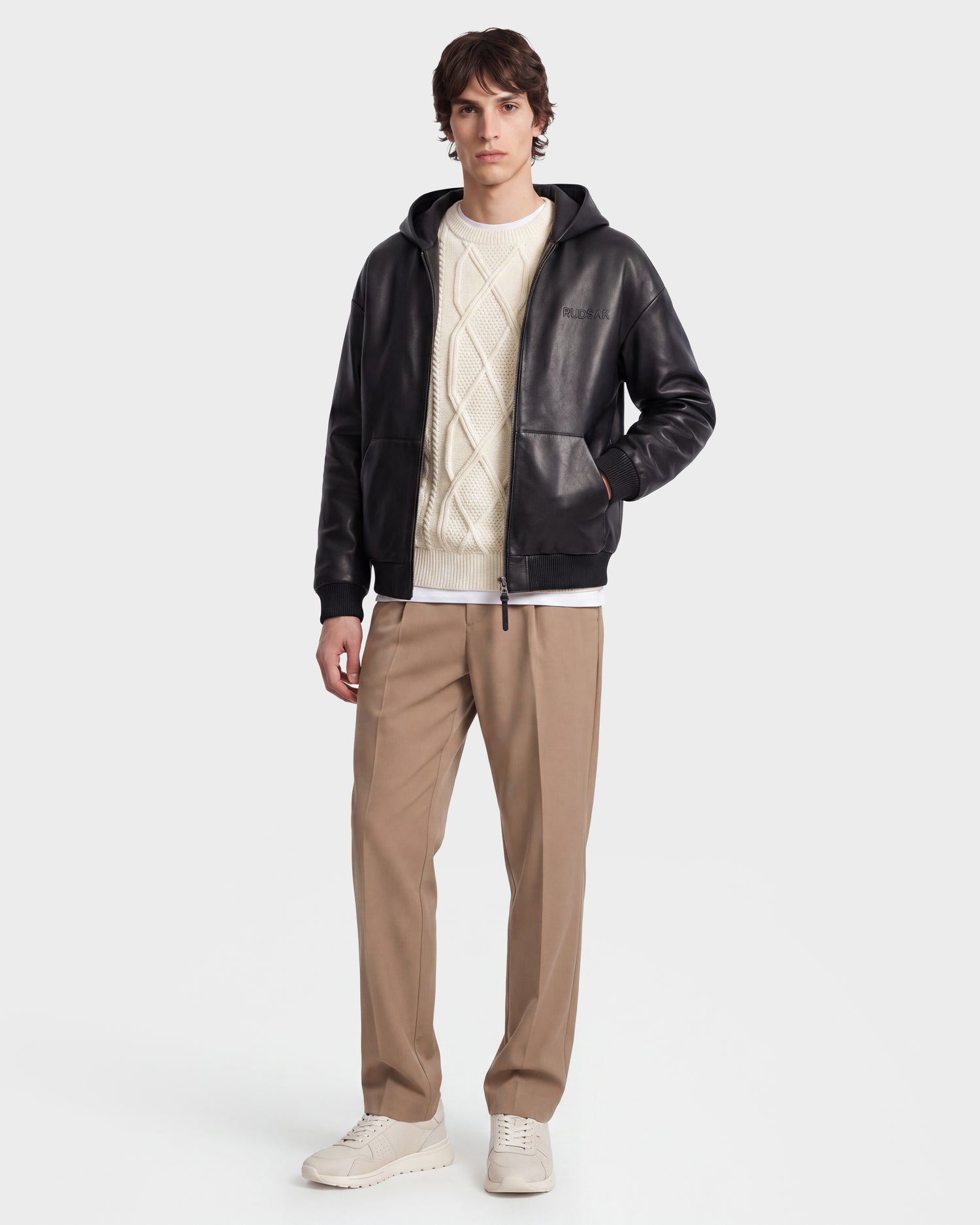 A man stands wearing the ARCHIE - 6126500 Black leather hoodie by Rudsak, layered over a white cable-knit sweater and styled with beige pants and white sneakers for an effortlessly urban look against a plain white background.