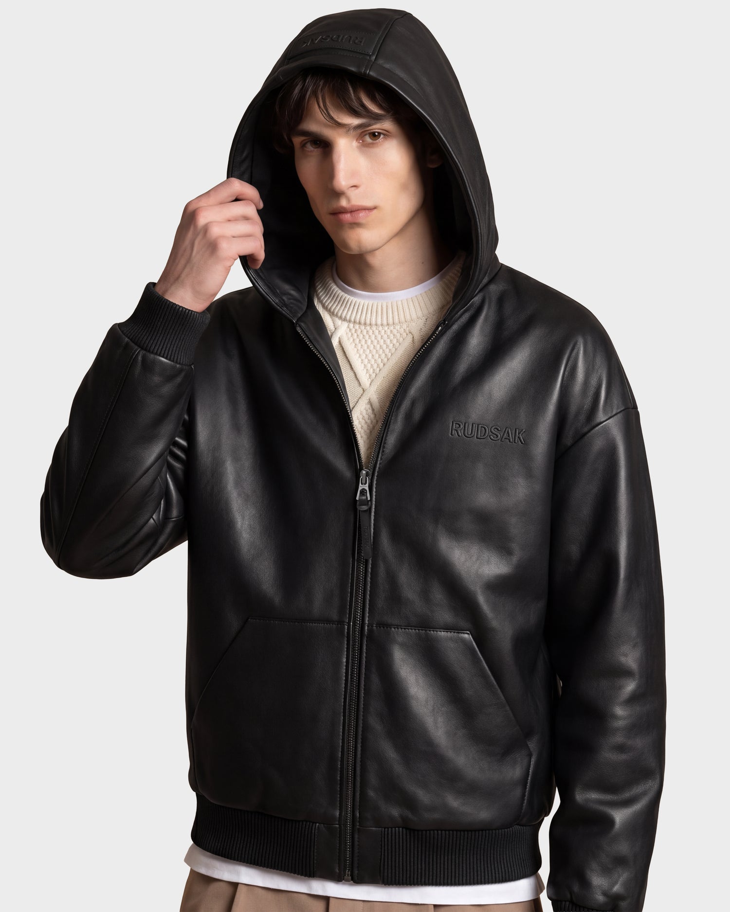 A young man wears the ARCHIE - 6126500 Black leather hoodie by Rudsak over a cream sweater, holding the hood with one hand. This urban look features a jacket with a front zipper, side pockets, and “RUDSAK” displayed on the chest, set against a neutral gray background.