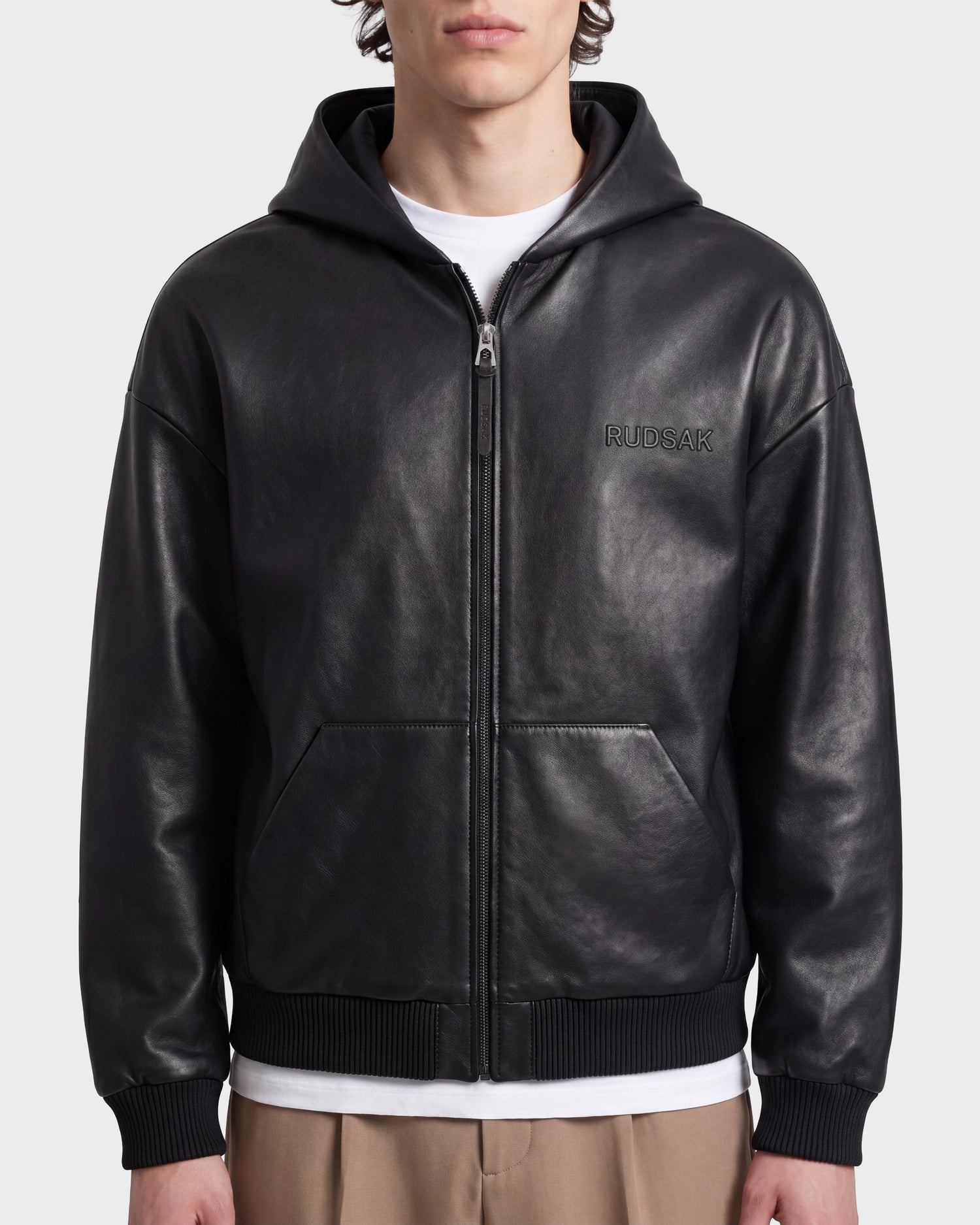 A person models urban style in the Rudsak ARCHIE - 6126500 Black leather hoodie, featuring the brand’s logo, layered over a white t-shirt and paired with beige pants. Posed against a plain background, the individual’s face is partly visible with a neutral expression.