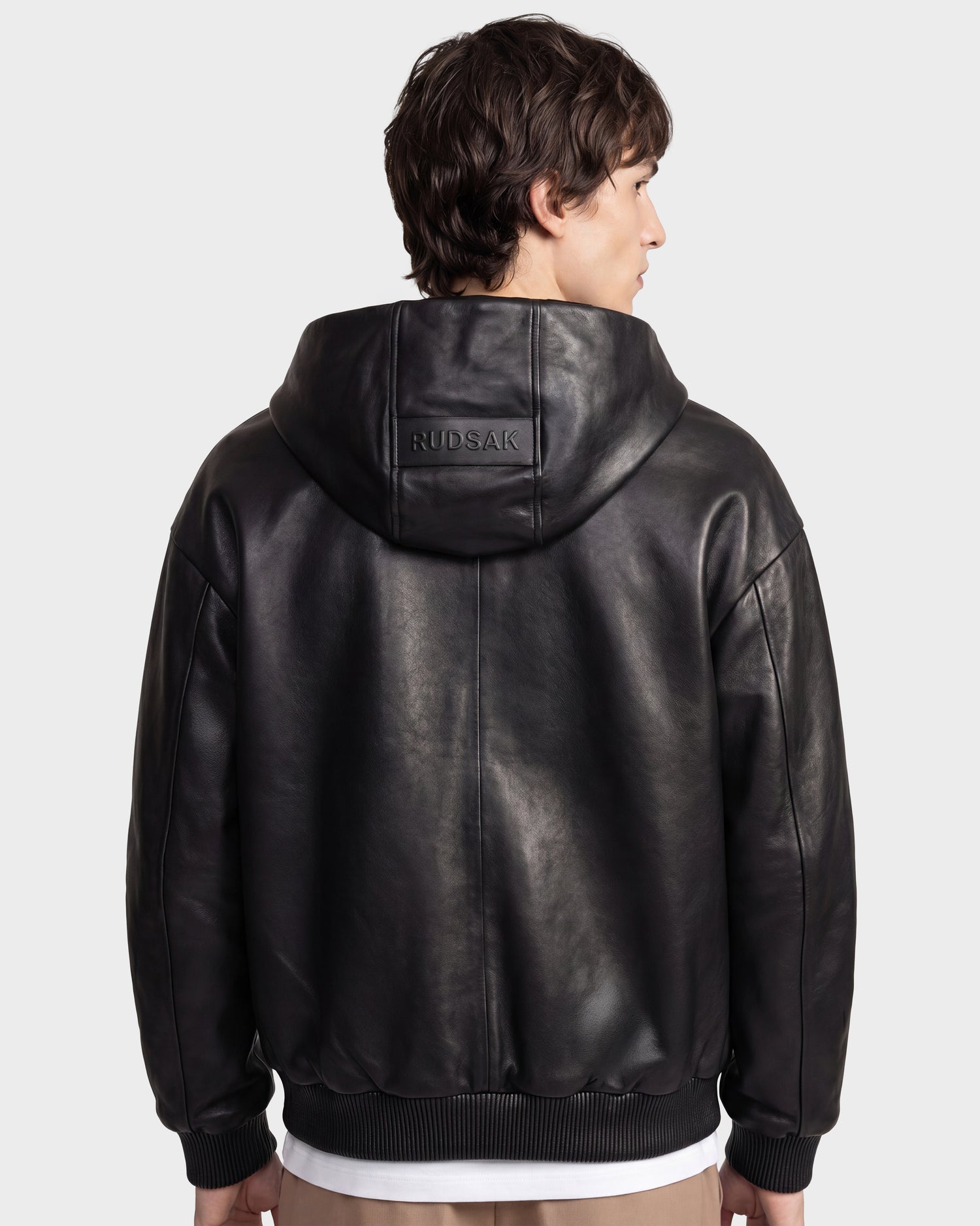 A person with short brown hair is shown from the back wearing the ARCHIE - 6126500 Black leather hoodie by Rudsak. The jacket, featuring urban styling, has a relaxed fit with ribbed cuffs and hem, and "RUDSAK" is embossed on the hood. The background is plain and light.