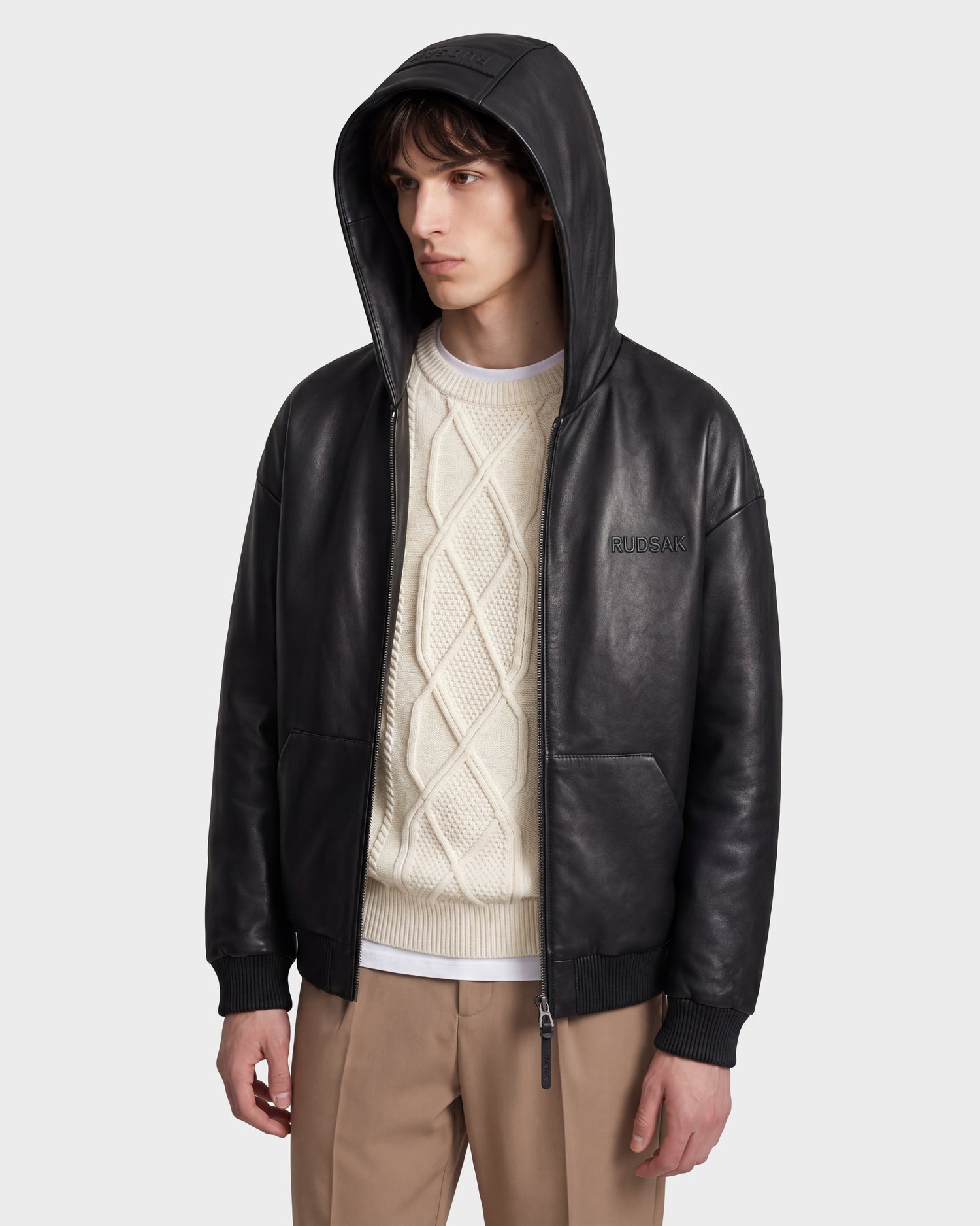 Wearing the Rudsak ARCHIE - 6126500 Black leather hoodie over a cream cable-knit sweater and tan pants, the individual stands against a plain light background, effortlessly showcasing urban style while glancing to the side.