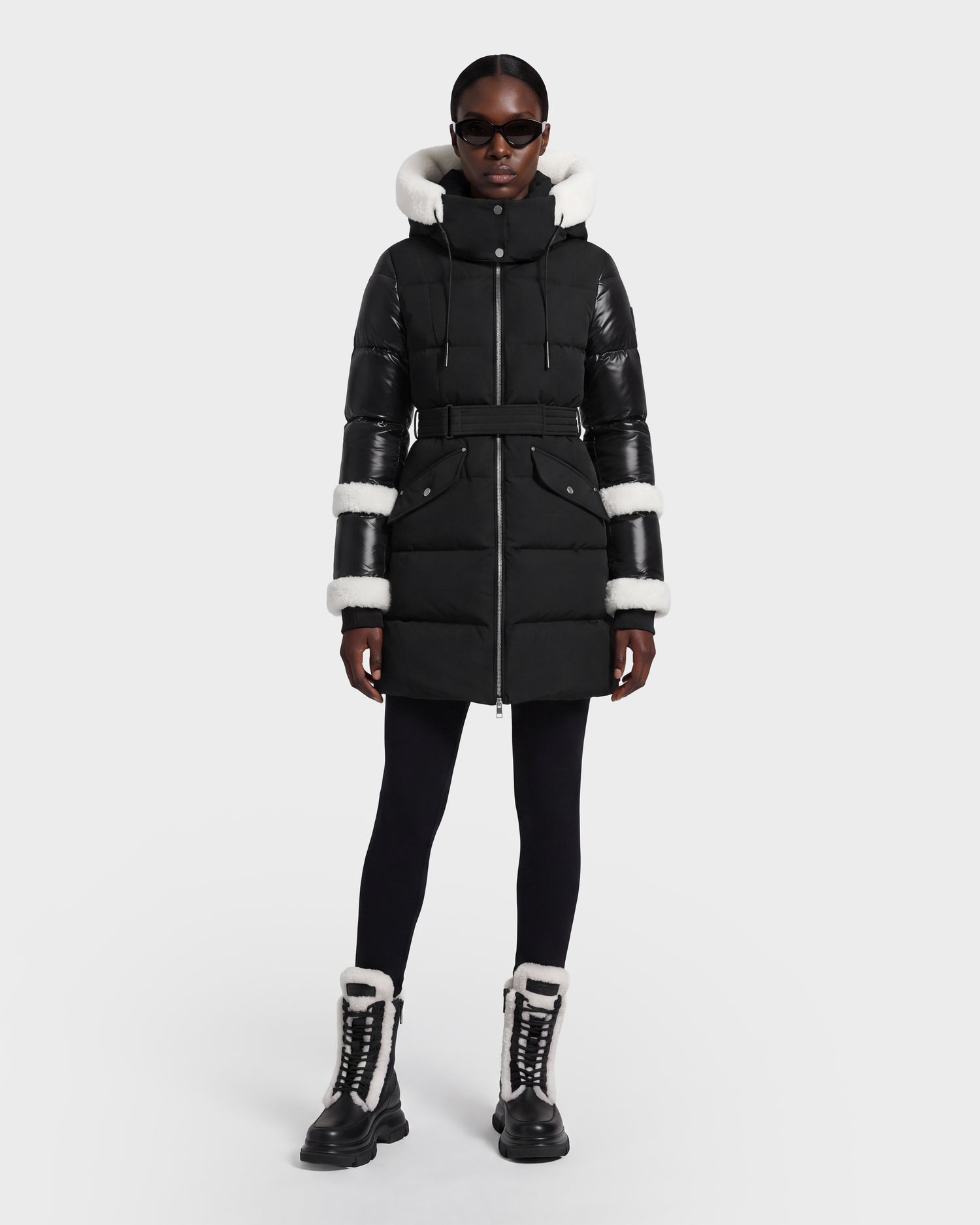 A person wearing the Rudsak AUDREY L - 8124585 Black/Iceland winter coat—featuring a black belted design crafted from recycled Stormshell fabric with white faux fur trim—paired with black leggings, black-and-white lace-up boots, and sunglasses stands facing forward against a plain white background.