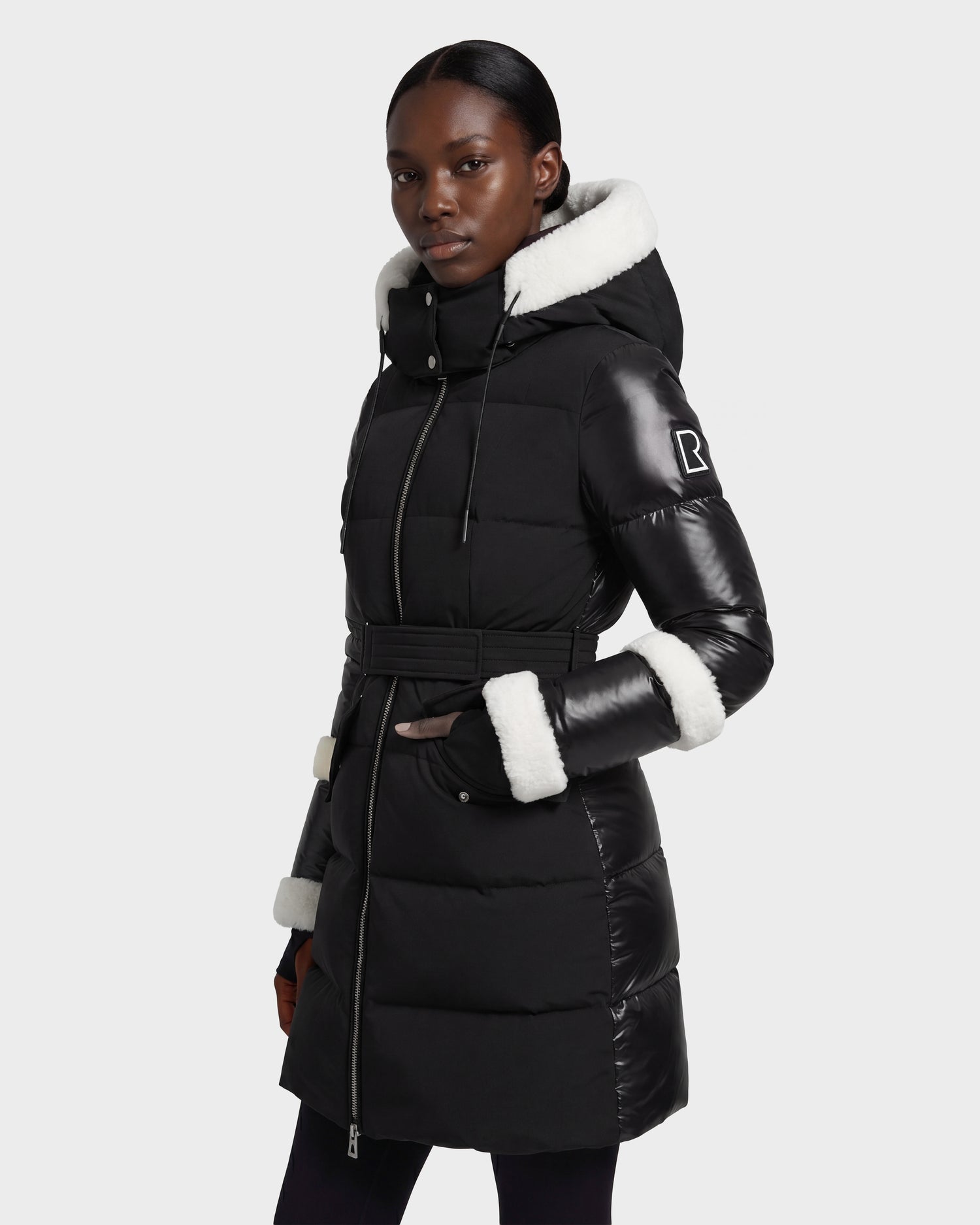 A woman stands confidently against a plain light background, wearing the Rudsak AUDREY L - 8124585 Black/Iceland down puffer jacket made from recycled Stormshell fabric, featuring white faux fur trim on the hood and cuffs.