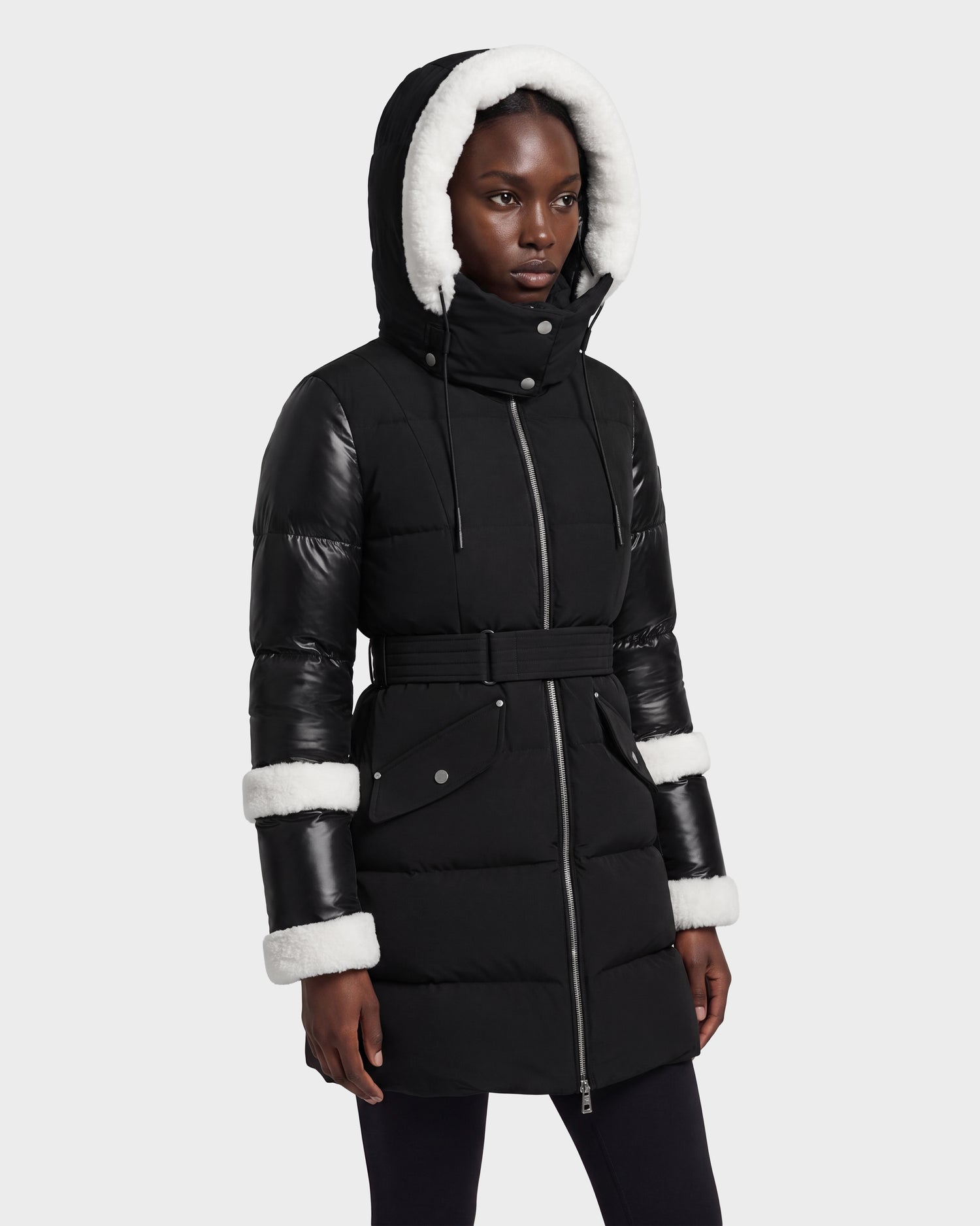 A woman wearing the Rudsak AUDREY L - 8124585 Black/Iceland coat, a black hooded winter coat made from recycled Stormshell fabric featuring white faux fur trim on the hood and cuffs, black leather sleeves, and a belted waist, stands against a plain light background.