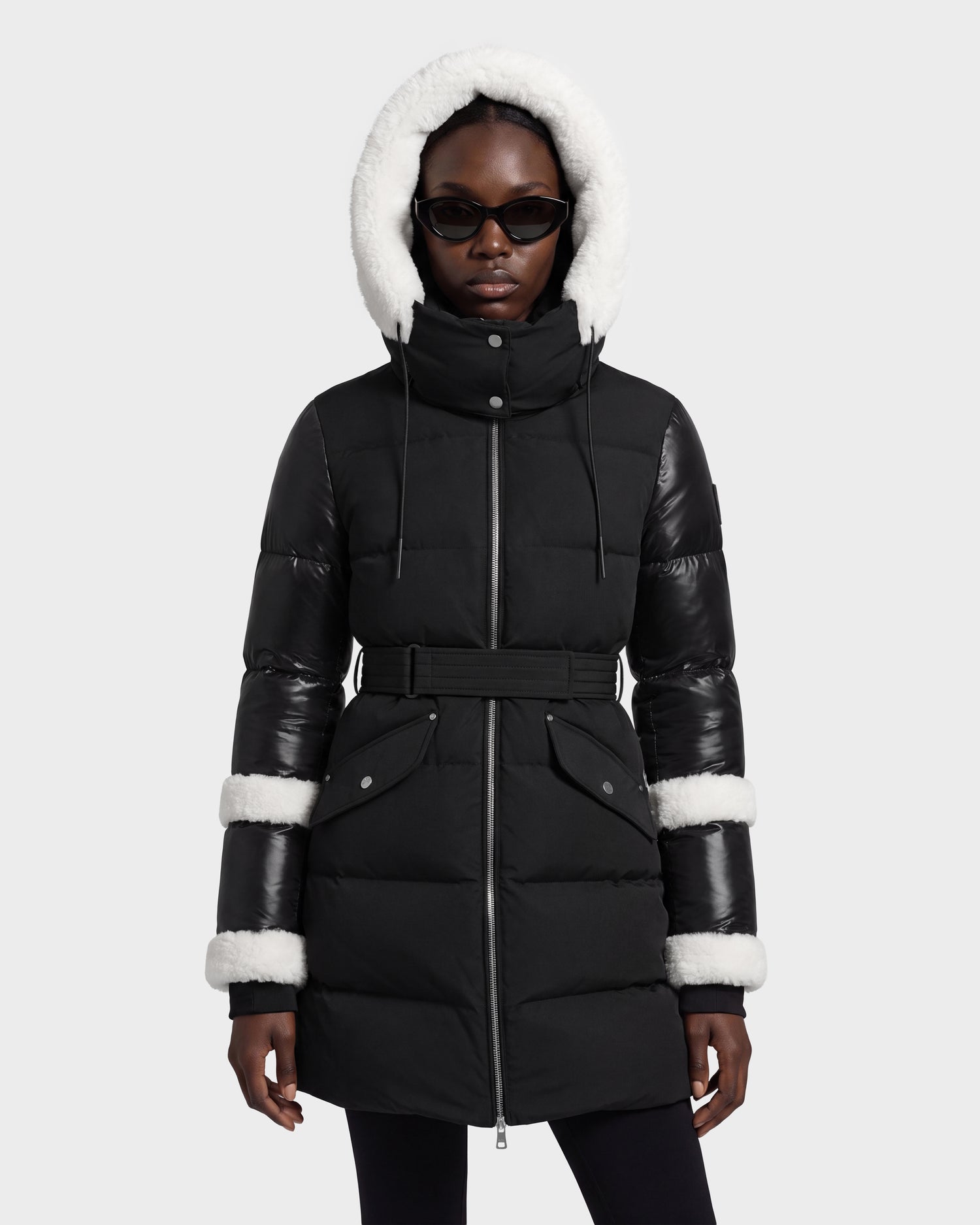 A person wearing the Rudsak AUDREY L - 8124585 Black/Iceland coat, a black belted winter jacket made from recycled Stormshell fabric with white faux fur trim on the hood and sleeves, paired with dark sunglasses, stands against a plain light background.