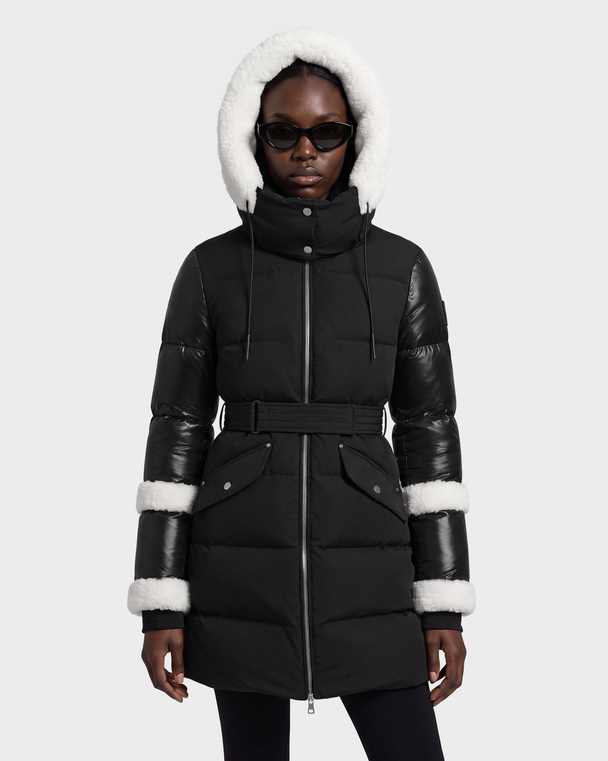 A person stands straight against a light gray background wearing the Rudsak AUDREY L - 8124585 Black/Iceland, a knee-length down puffer jacket featuring a white fur-lined hood, leather sleeve trim, black belt, and black sunglasses. The coat is crafted from recycled Stormshell fabric and insulated with RDS certified down for warmth and sustainability.