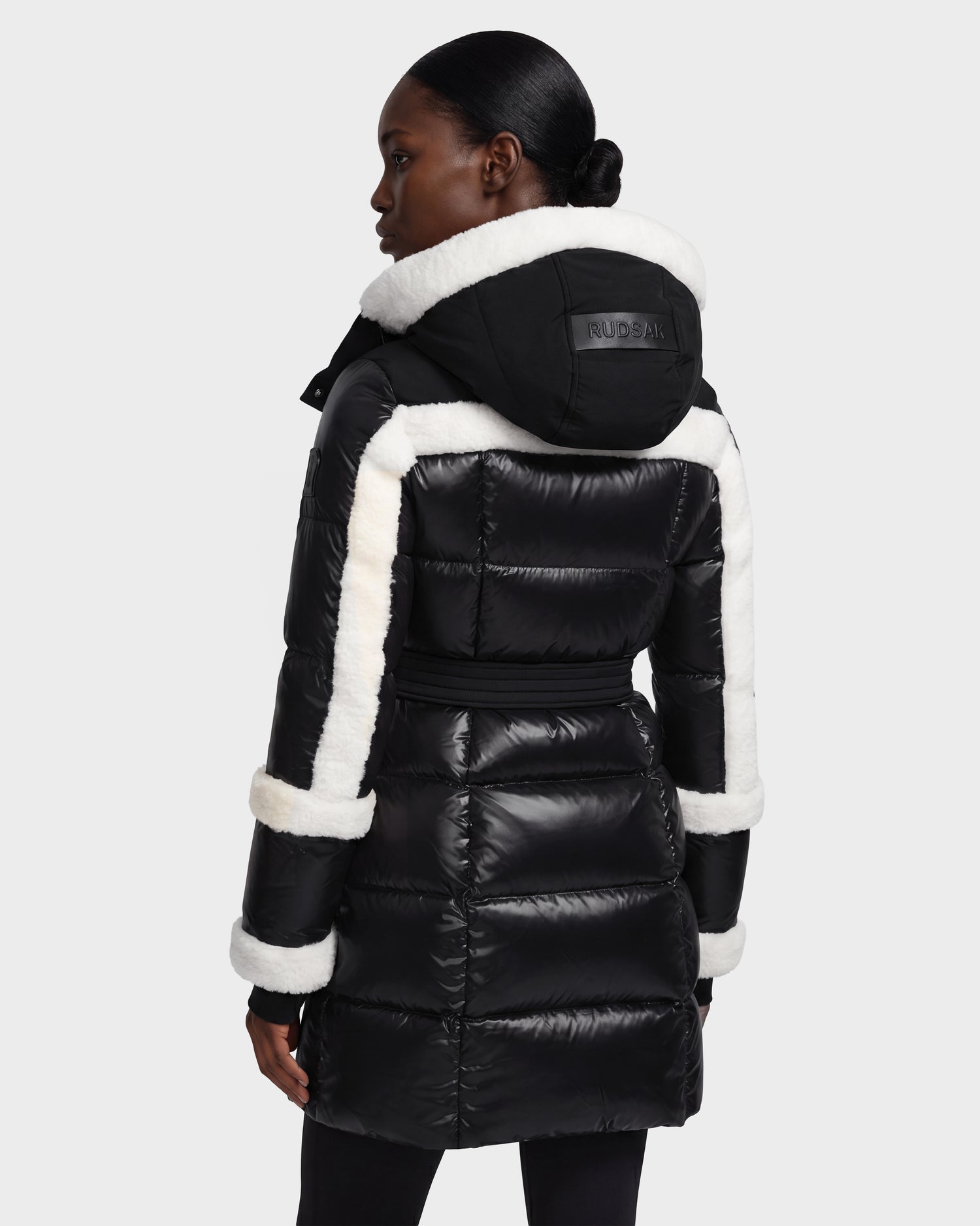 A woman is shown from the back against a plain light background, wearing the Rudsak AUDREY L - 8124585 Black/Iceland down puffer jacket—crafted from recycled Stormshell fabric—with shiny black finish and white fur trim along the hood, sleeves, and seams.