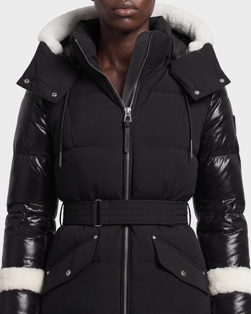 A person wearing the Rudsak AUDREY L - 8124585 Black/Iceland puffer jacket, a black belted down coat crafted from recycled Stormshell fabric, featuring shiny sleeves, a front zipper, snap pockets, and a hood lined with white fur trim.