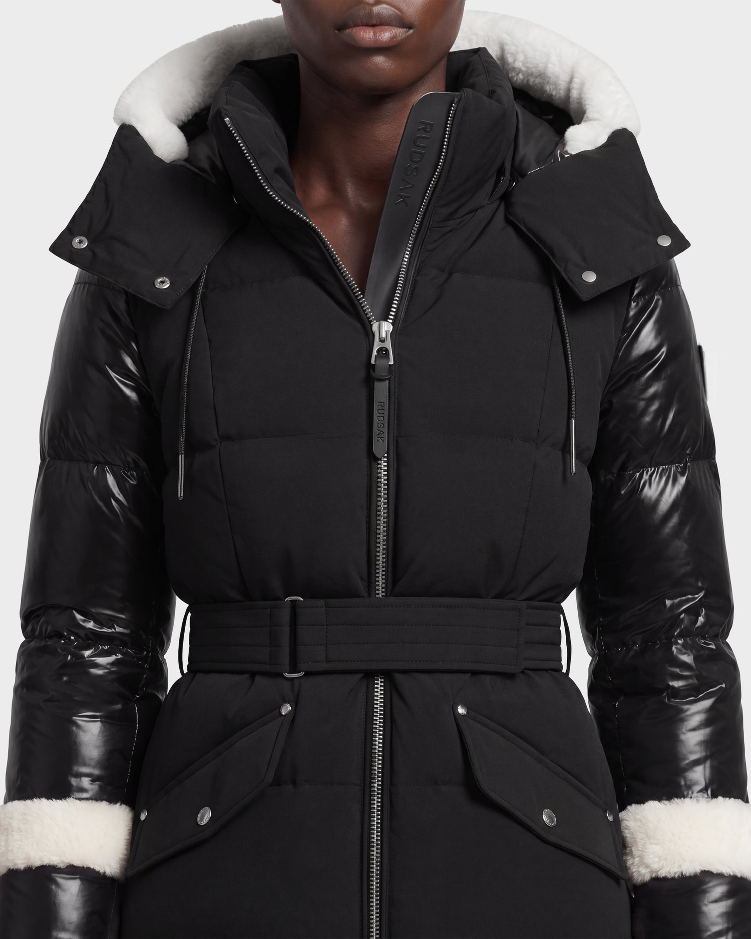 A person wearing the Rudsak AUDREY L - 8124585 Black/Iceland belted down puffer jacket, featuring shiny sleeves, white shearling trim on the hood and cuffs, and large front pockets, crafted from recycled Stormshell fabric and shown from shoulders to waist.