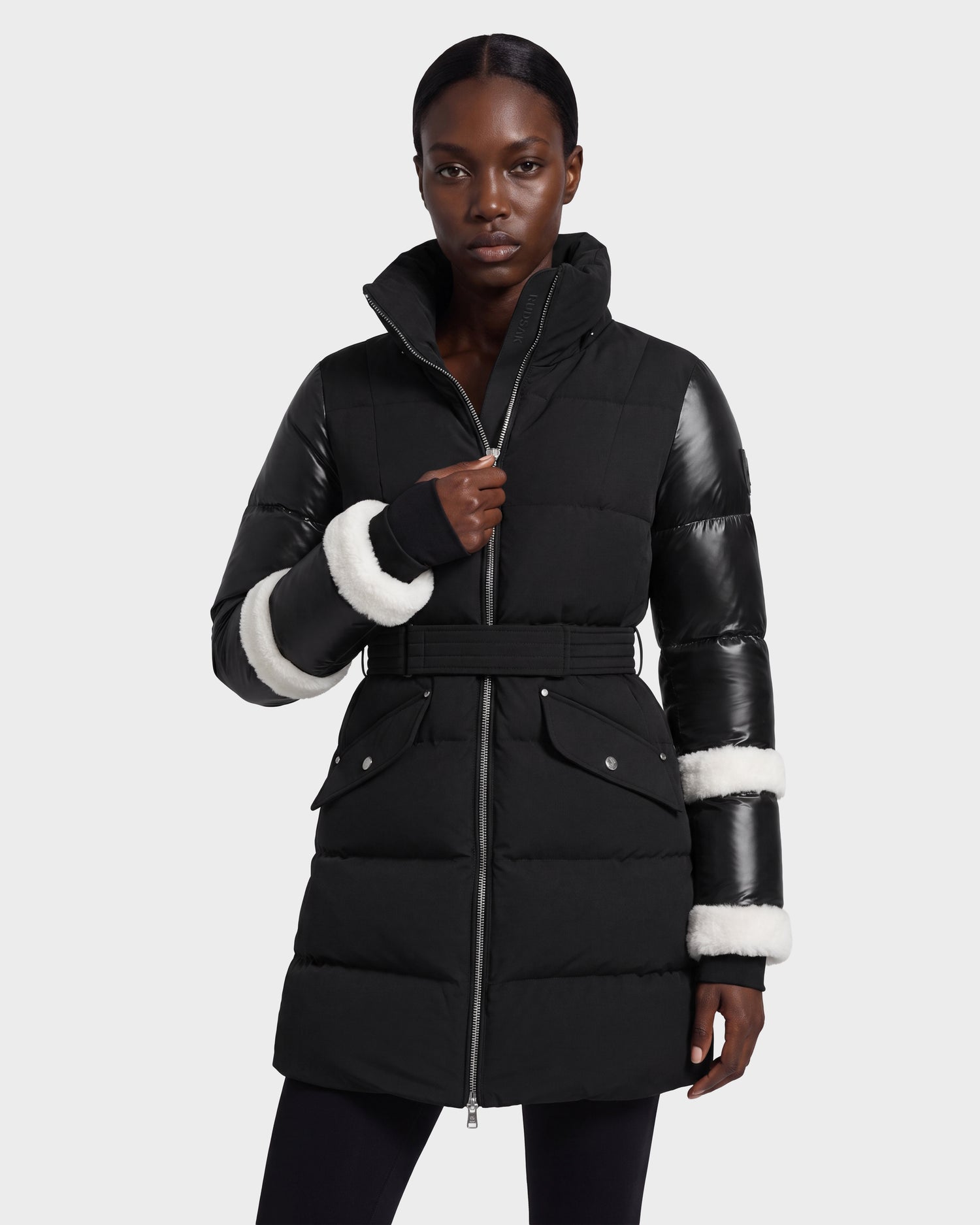 A person stands against a plain light background wearing the Rudsak AUDREY L - 8124585 Black/Iceland puffer jacket. This black, belted down coat features leather sleeves with white fur trim, front flap pockets, and is crafted from recycled Stormshell fabric for both style and sustainability.