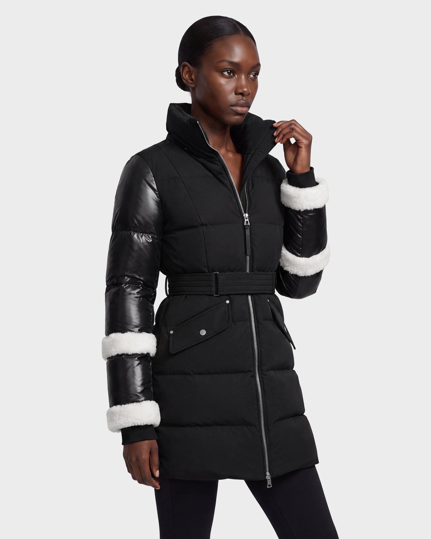 A woman stands against a plain light background wearing the Rudsak AUDREY L - 8124585 Black/Iceland, a knee-length down puffer jacket crafted from recycled Stormshell fabric, featuring shiny black sleeves and white faux fur trim on the cuffs.