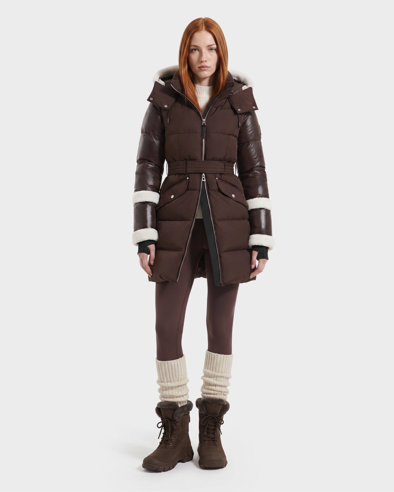A woman with straight red hair wears the Rudsak AUDREY L - 8124585 Espresso knee-length down puffer jacket, crafted from recycled Stormshell fabric with white trim, paired with brown leggings, leg warmers, and brown boots, standing against a plain white background.
