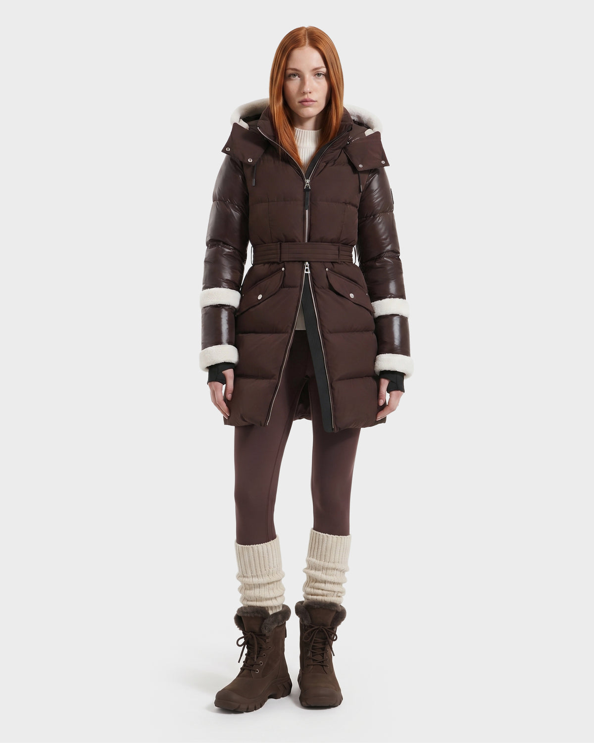 A woman with straight red hair models the Rudsak AUDREY L - 8124585 Espresso, a knee-length down puffer jacket crafted from recycled Stormshell fabric and filled with RDS-certified down. The jacket features rich brown tones and a white fur trim. She pairs it with brown leggings, tall slouchy socks, and brown boots while standing against a plain white background.