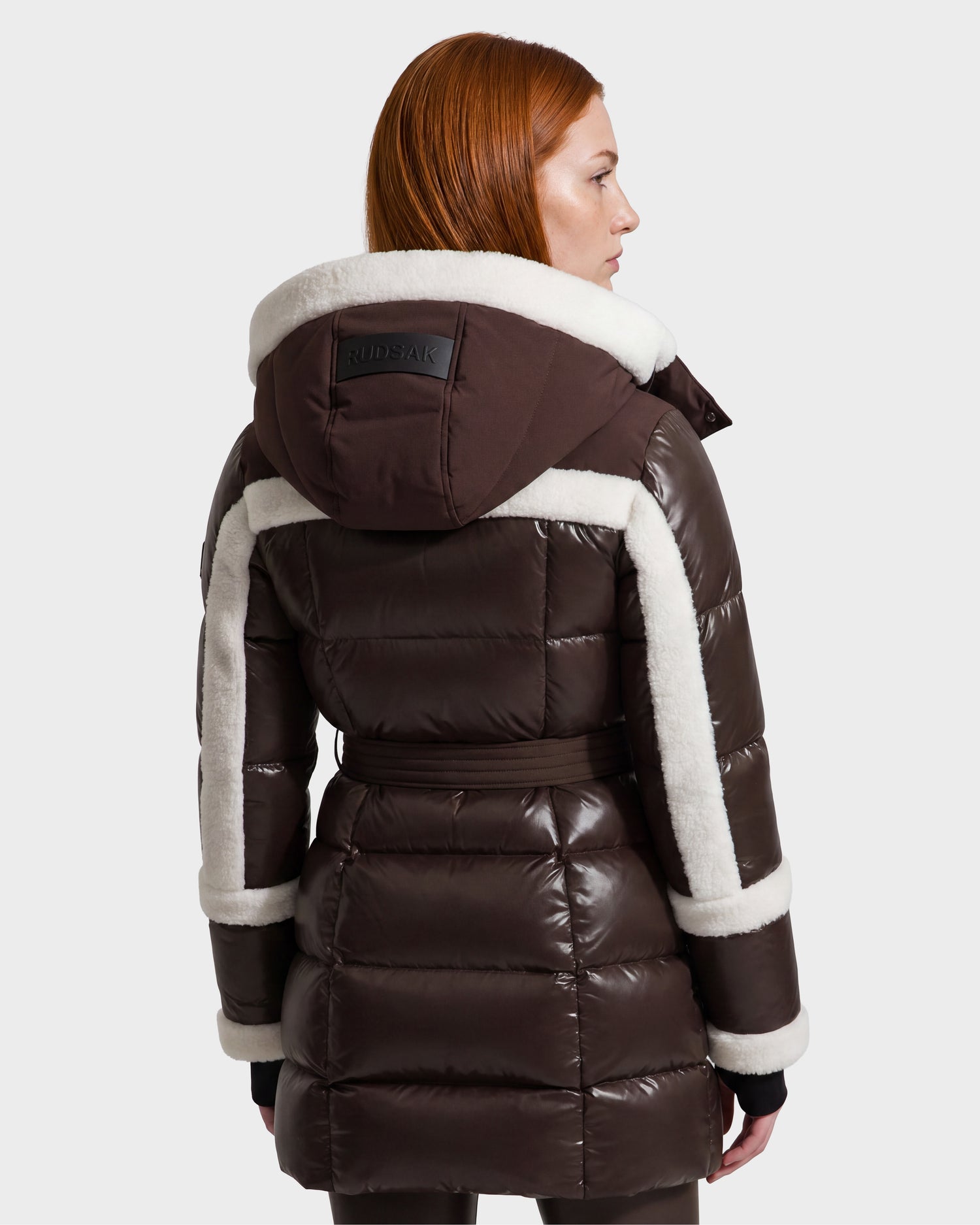 A person with straight red hair is seen from the back, wearing the Rudsak AUDREY L - 8124585 Espresso knee-length down puffer jacket, crafted from recycled Stormshell fabric in a dark brown hue, featuring white shearling trim and a hood, set against a plain light background.