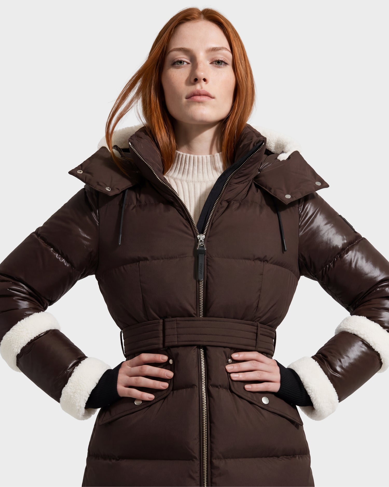 A woman with long red hair stands against a light gray background, wearing the Rudsak AUDREY L - 8124585 Espresso knee-length down puffer jacket. The jacket is crafted from recycled Stormshell fabric and features a brown, belted silhouette with white fur trim on the hood and cuffs.