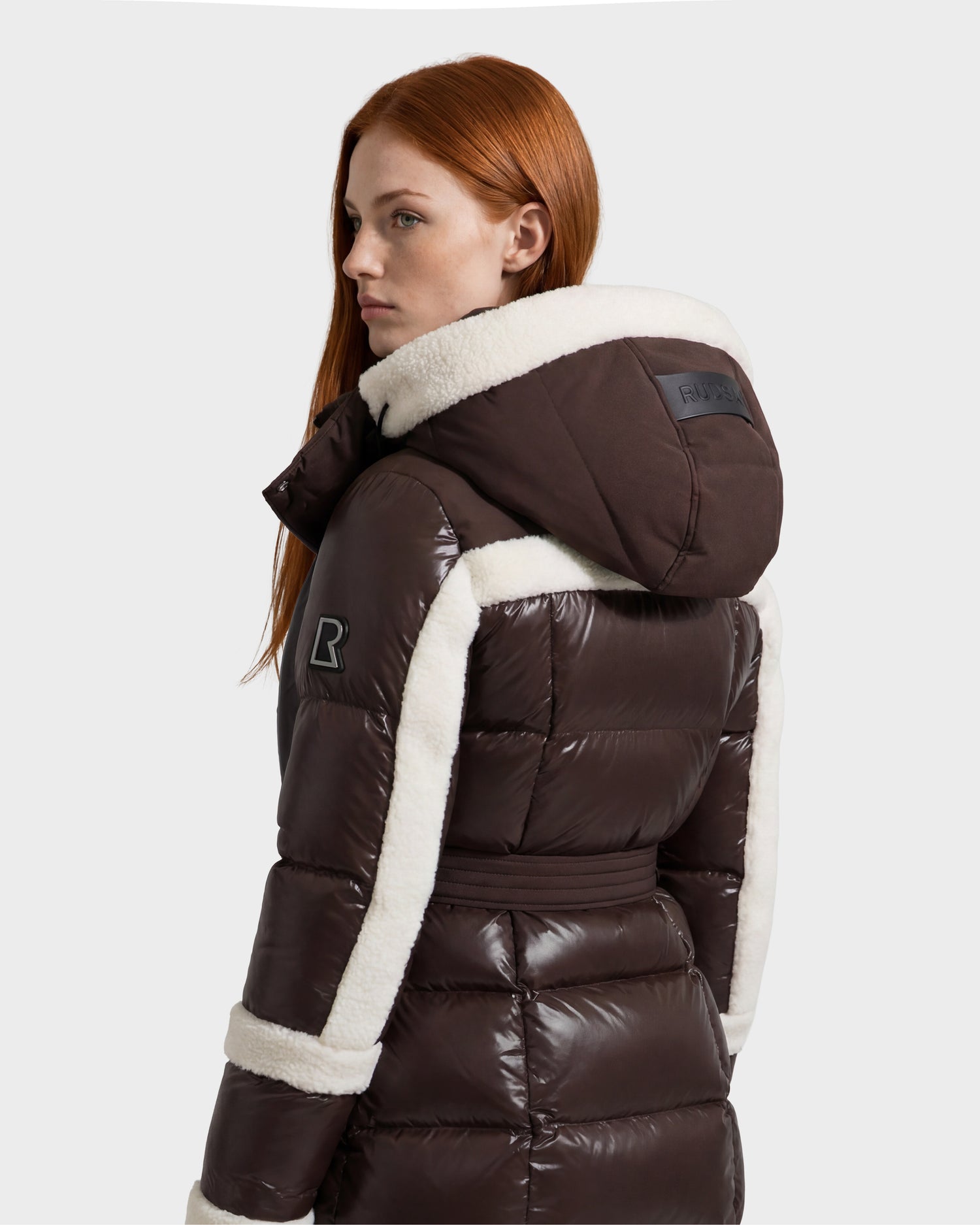 A woman with long red hair wears the Rudsak AUDREY L - 8124585 Espresso, a glossy brown knee-length down puffer jacket featuring white sherpa trim and a hood. Crafted from recycled Stormshell fabric and filled with RDS certified down, she stands looking to the side against a plain light background.