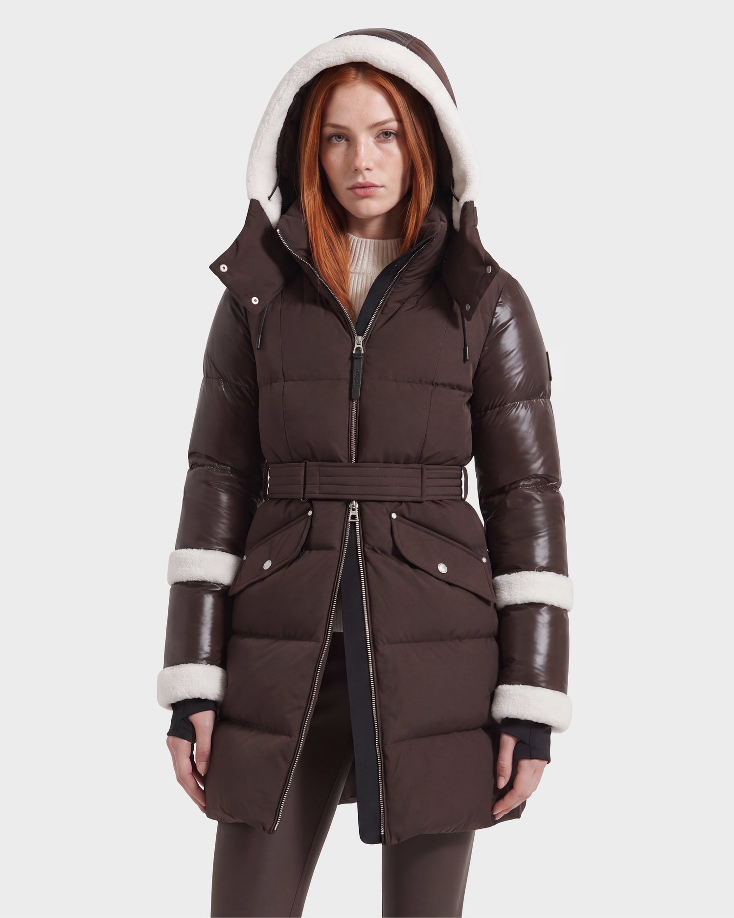 A woman with long red hair wears the Rudsak AUDREY L – 8124585 Espresso, a knee-length down puffer jacket in brown. The coat features a belted waist, white faux fur trim on the hood and cuffs, and is crafted from recycled Stormshell fabric filled with RDS certified down. She stands against a plain light background.