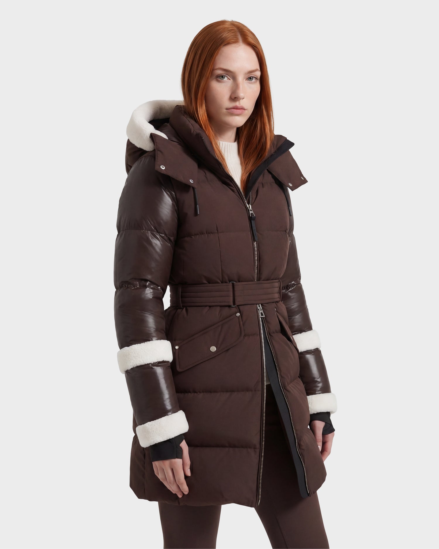 A woman with long red hair models the Rudsak AUDREY L - 8124585 Espresso, a dark brown, knee-length down puffer jacket made from recycled Stormshell fabric. The jacket features a hood, belted waist, and white trim on the cuffs, set against a light grey background.