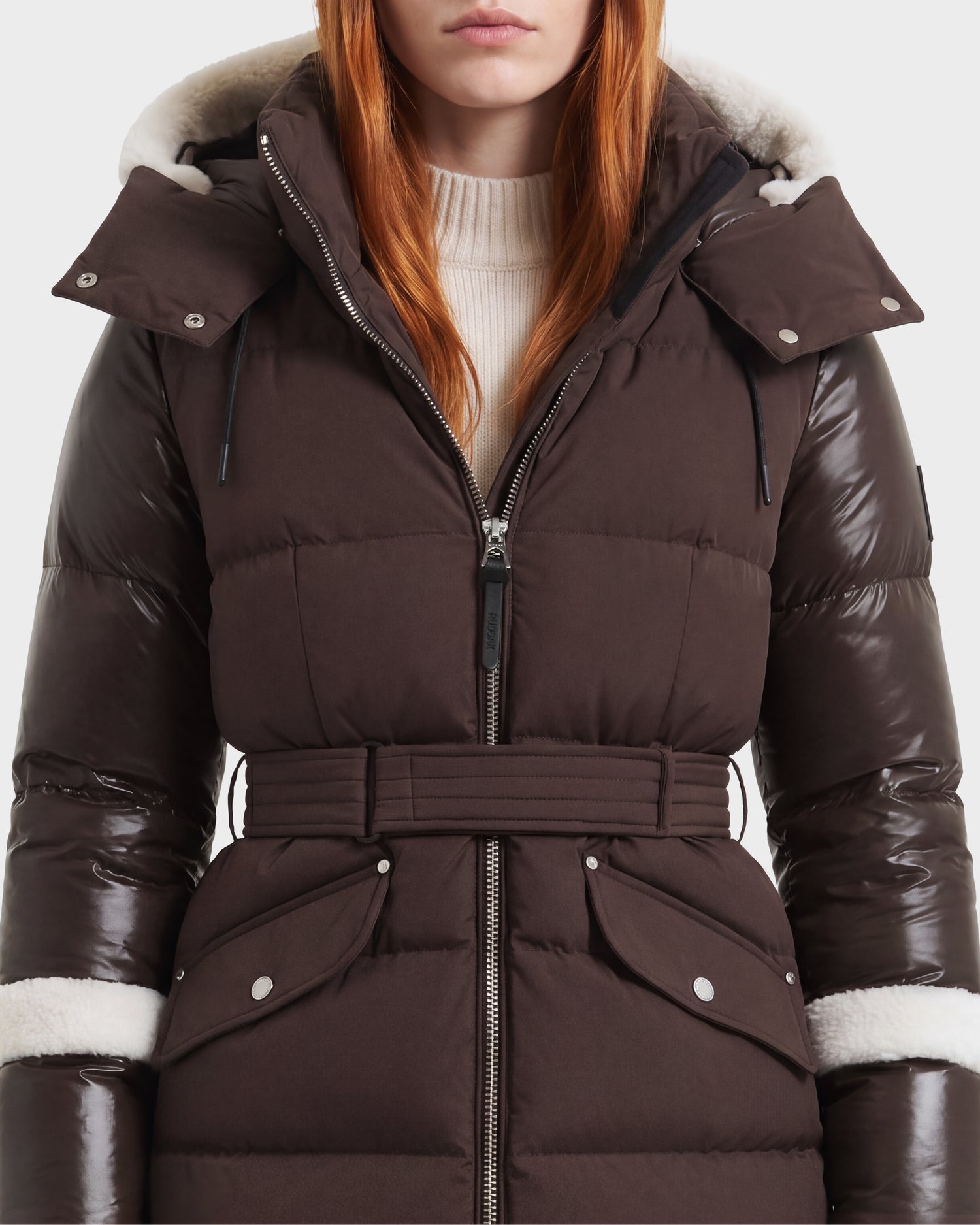 A woman wears the Rudsak AUDREY L - 8124585 Espresso, a knee-length down puffer coat made from recycled Stormshell fabric. The jacket features a belted waist, shiny sleeves, fur trim, and a hood, layered over a cream turtleneck sweater. Filled with RDS-certified down, the dark espresso brown coat complements her long red hair. The image is cropped at her nose and mid-thigh.