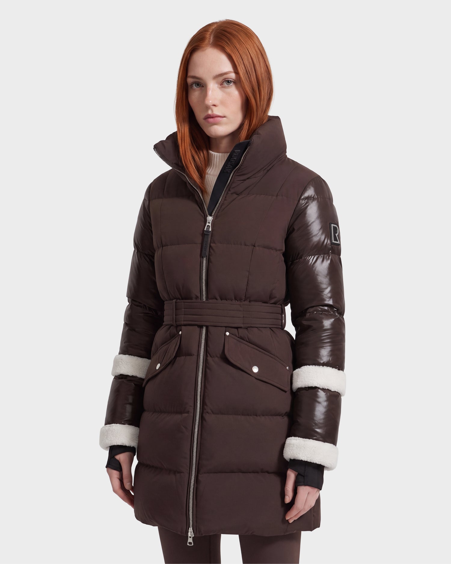A woman with straight red hair models the Rudsak AUDREY L - 8124585 down puffer jacket in Espresso, a knee-length coat featuring shiny sleeves, white trim, and a belted waist. Crafted from recycled Stormshell fabric and filled with RDS certified down, she stands facing forward against a plain background.