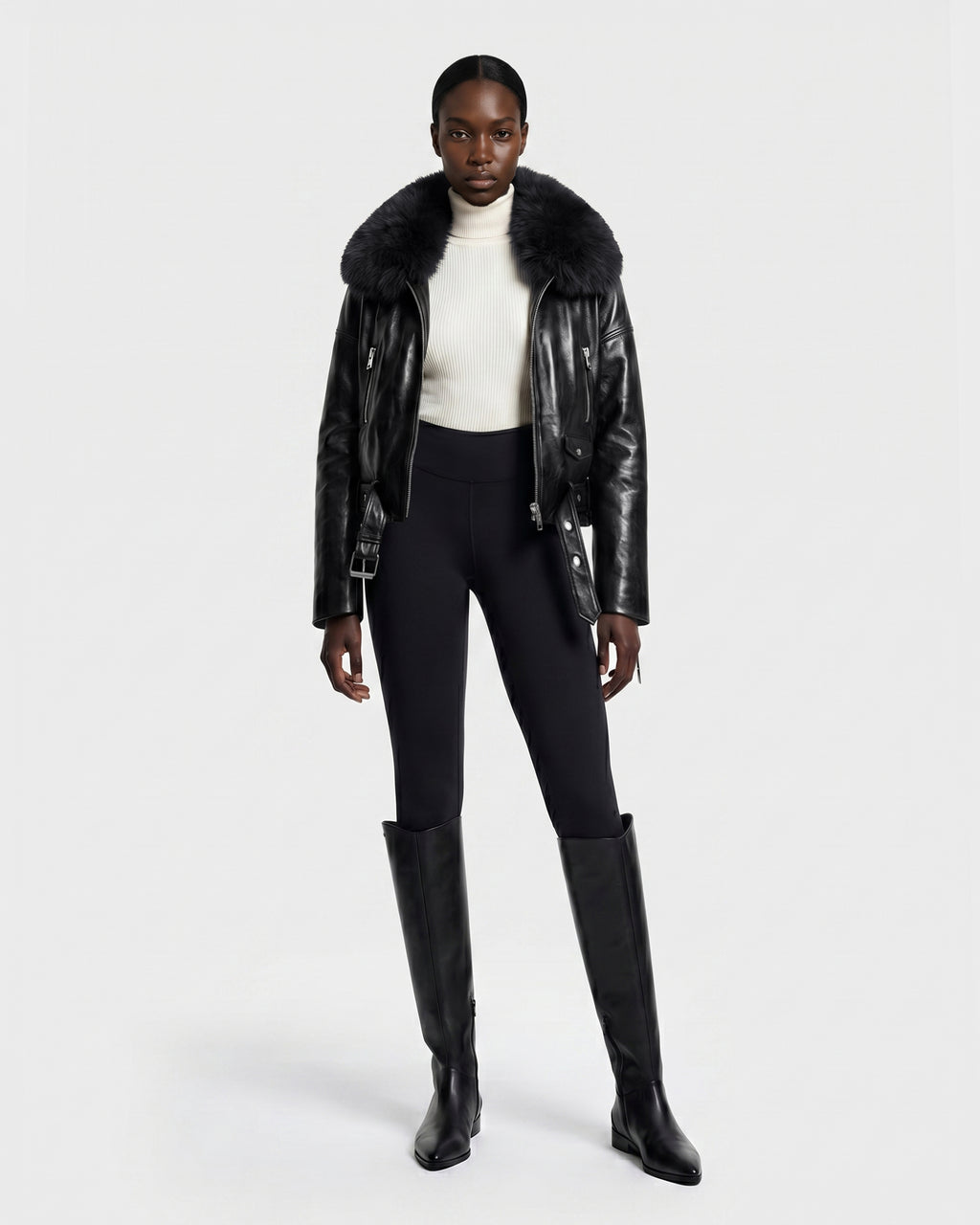 A woman stands against a plain white background wearing the Rudsak AVITA - 8126938 Black leather jacket with a fur collar, paired with a white turtleneck sweater, black fitted pants, and tall black boots.