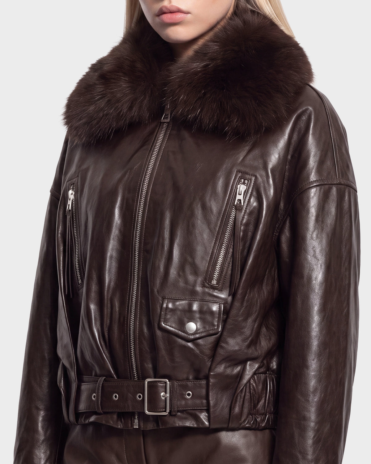 A woman models the Rudsak AVITA - 8126938 Espresso jacket, a dark brown leather piece featuring a fur collar, zippered chest pockets, a belted waist, and metallic snap accents. The image is cropped above her mouth and at the hips.