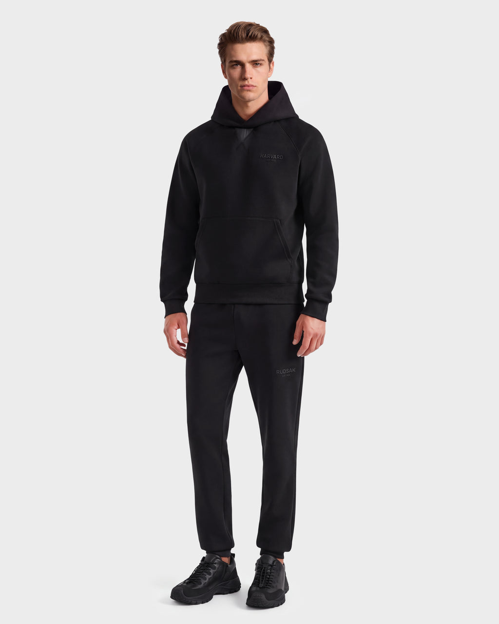 A man stands facing forward, wearing the limited-edition Rudsak BARRY HV - H525500 Black hooded sweatshirt, paired with matching black jogger pants and black sneakers, against a plain light background.