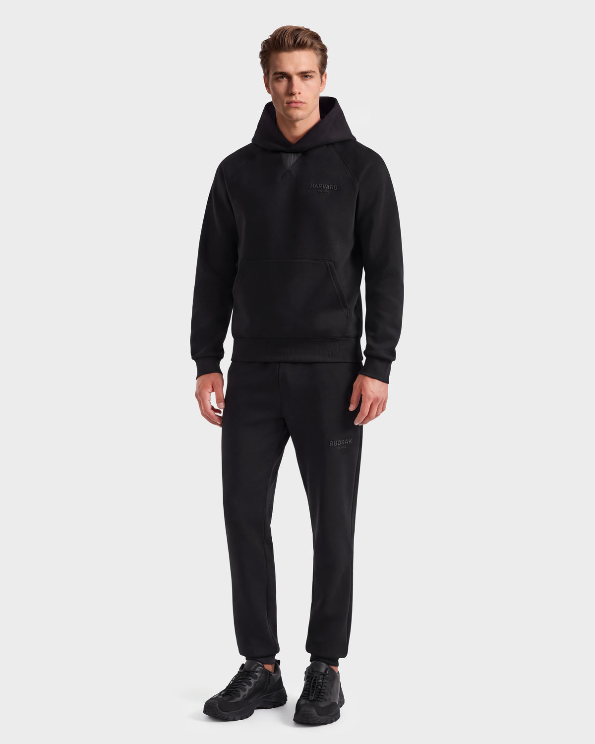 A man stands facing forward, wearing the limited-edition Rudsak BARRY HV - H525500 Black hooded sweatshirt, paired with matching black jogger pants and black sneakers, against a plain light background.