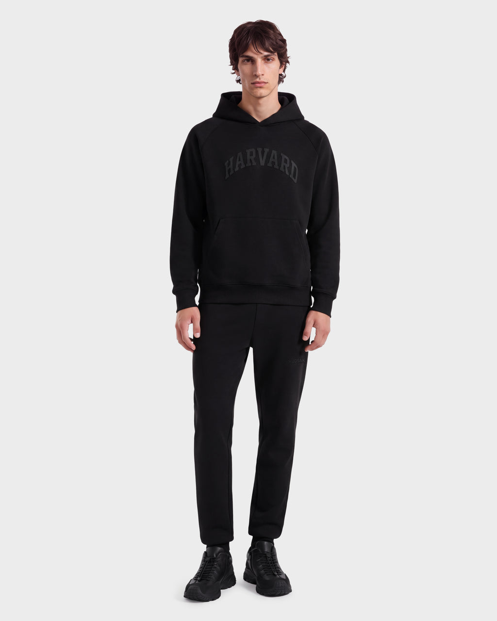 A man stands facing forward, wearing the Rudsak BRIAR R HV - H525506 Black limited-edition hoodie with matching black pants and black shoes against a plain light gray background.