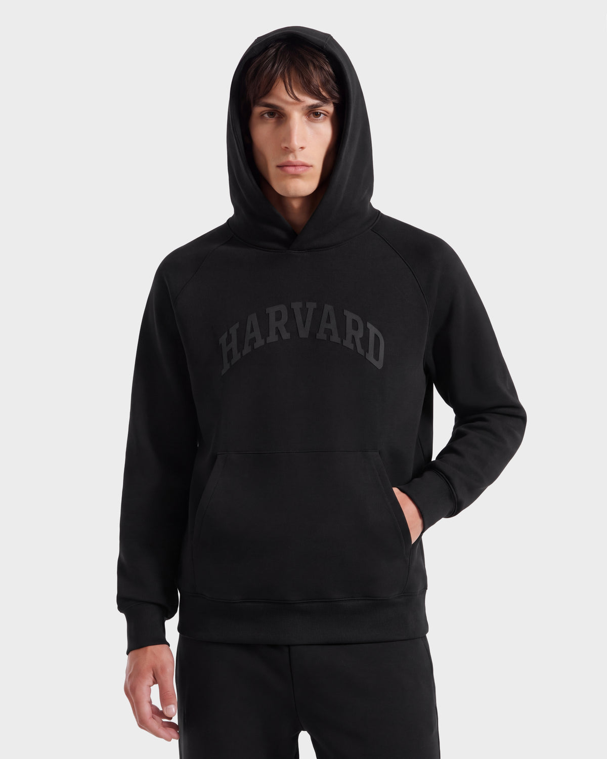 A person wearing the Rudsak BRIAR R HV - H525506 Black hoodie, featuring “HARVARD” on the front, stands against a plain light background with one hand in the hoodie pocket.