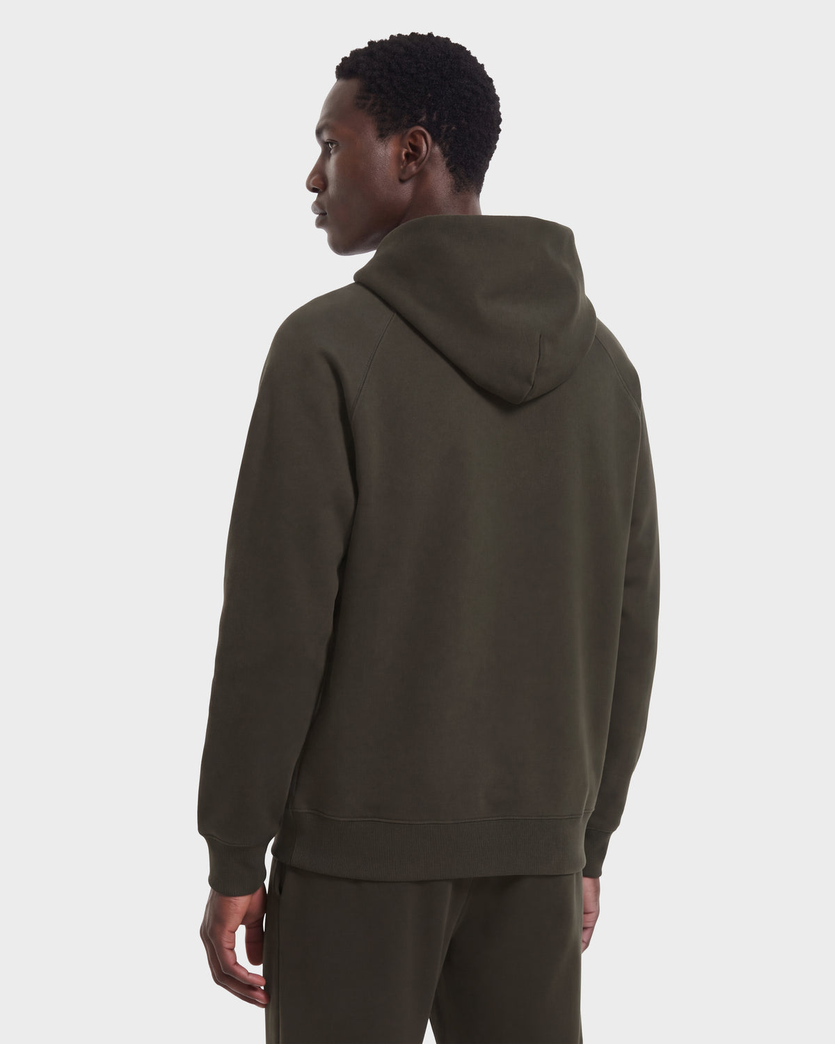 A person wearing the Rudsak BRIAR R HV - H525506 Olive limited-edition dark green hooded sweatshirt and matching pants stands facing away, looking to the side, against a plain white background.