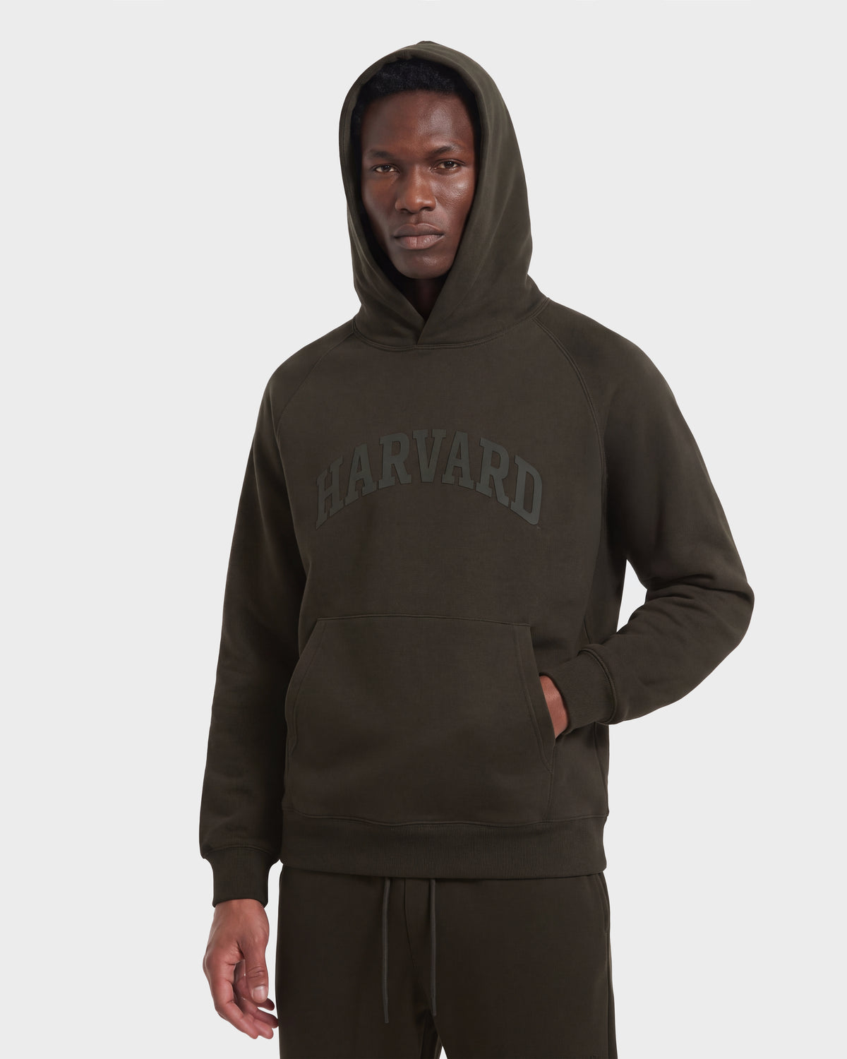 A person is wearing the Rudsak BRIAR R HV - H525506 Olive hoodie, a dark green limited-edition piece, featuring “Harvard” printed on the front. They are standing facing forward against a plain white background, with one hand in the hoodie pocket.