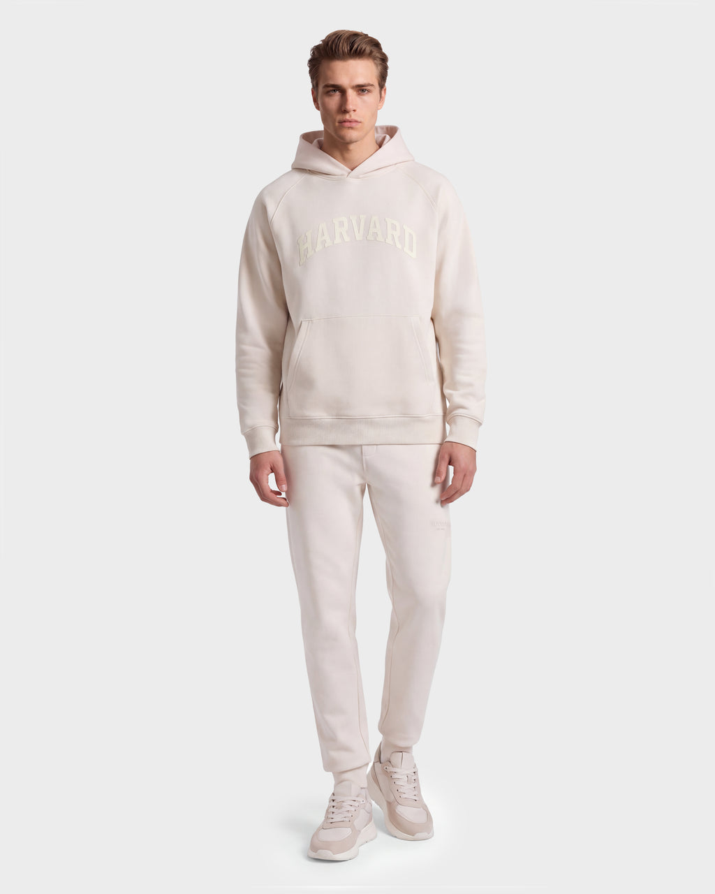 A man stands facing forward, wearing a Rudsak BRIAR R HV - H525506 hoodie in Vanilla, with matching sweatpants and sneakers. The plain, light background emphasizes his casual outfit.