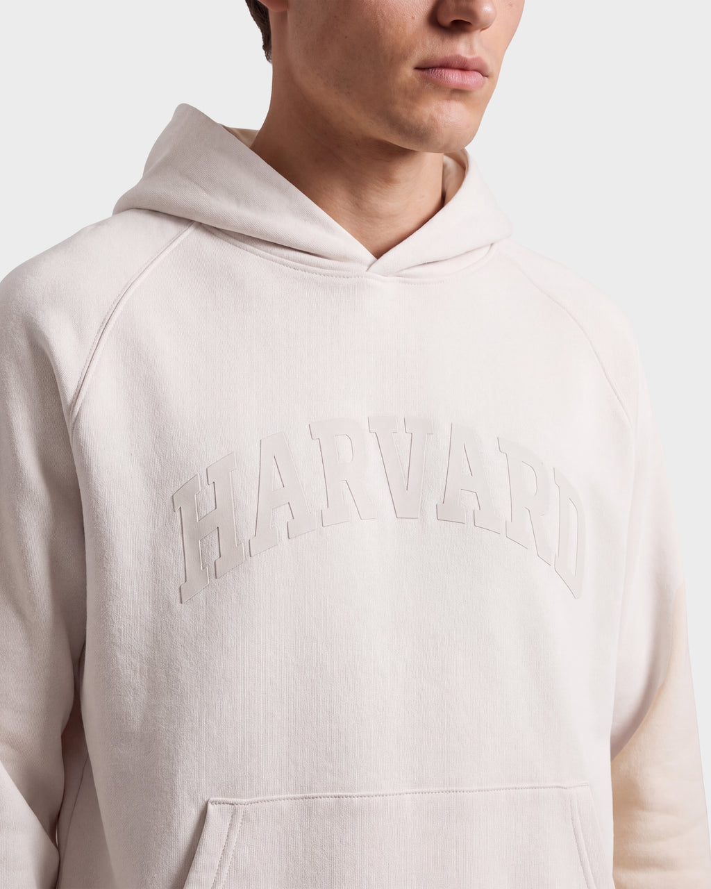 A person is seen wearing the Rudsak BRIAR R HV - H525506 Vanilla hoodie, featuring the word HARVARD embossed on the front. The image highlights classic collegiate style and focuses on the upper torso and part of the face against a plain background.
