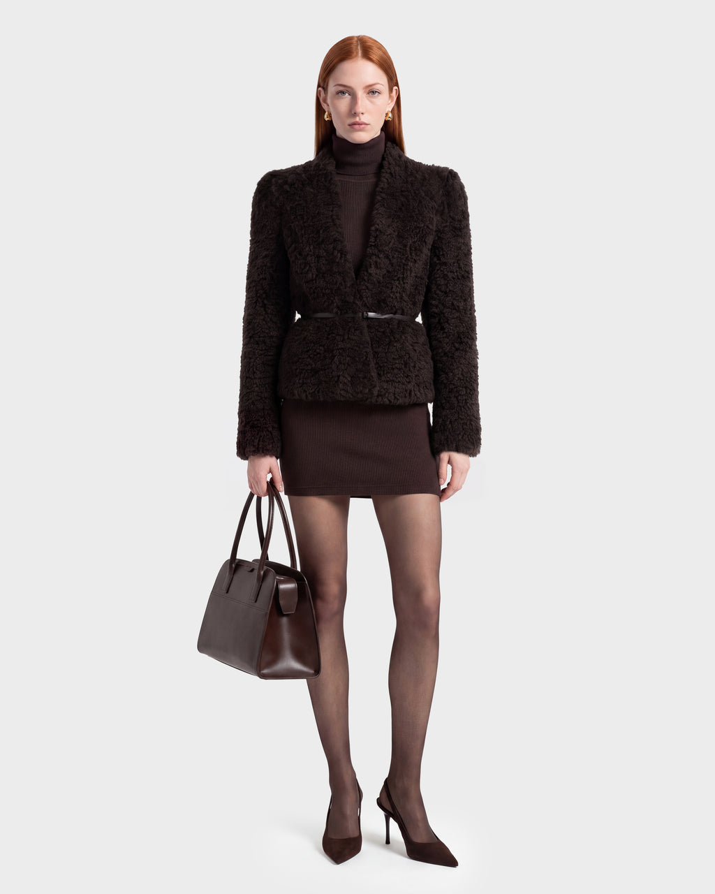 A woman with long red hair wears the Rudsak BRINLEY - 8126980 Espresso fuzzy jacket, a matching turtleneck and mini skirt, sheer tights, and heels. She holds a dark brown handbag and stands against a plain, light background.