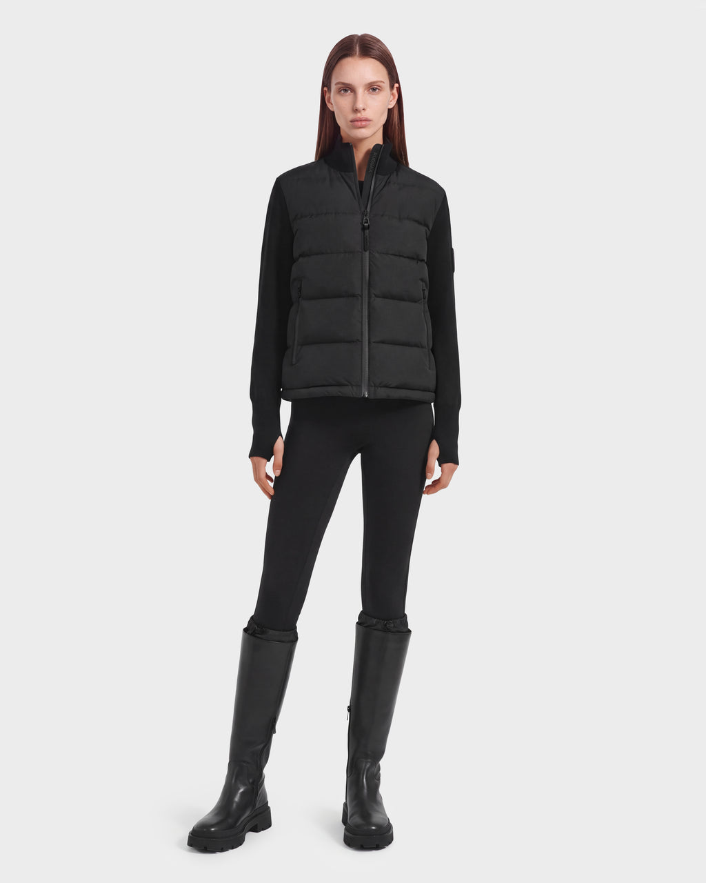 A person stands facing forward against a plain white background, wearing the limited-edition Rudsak CAMILLE HV - H125500 Black down jacket layered over a black long-sleeve top, black leggings, and tall black boots.