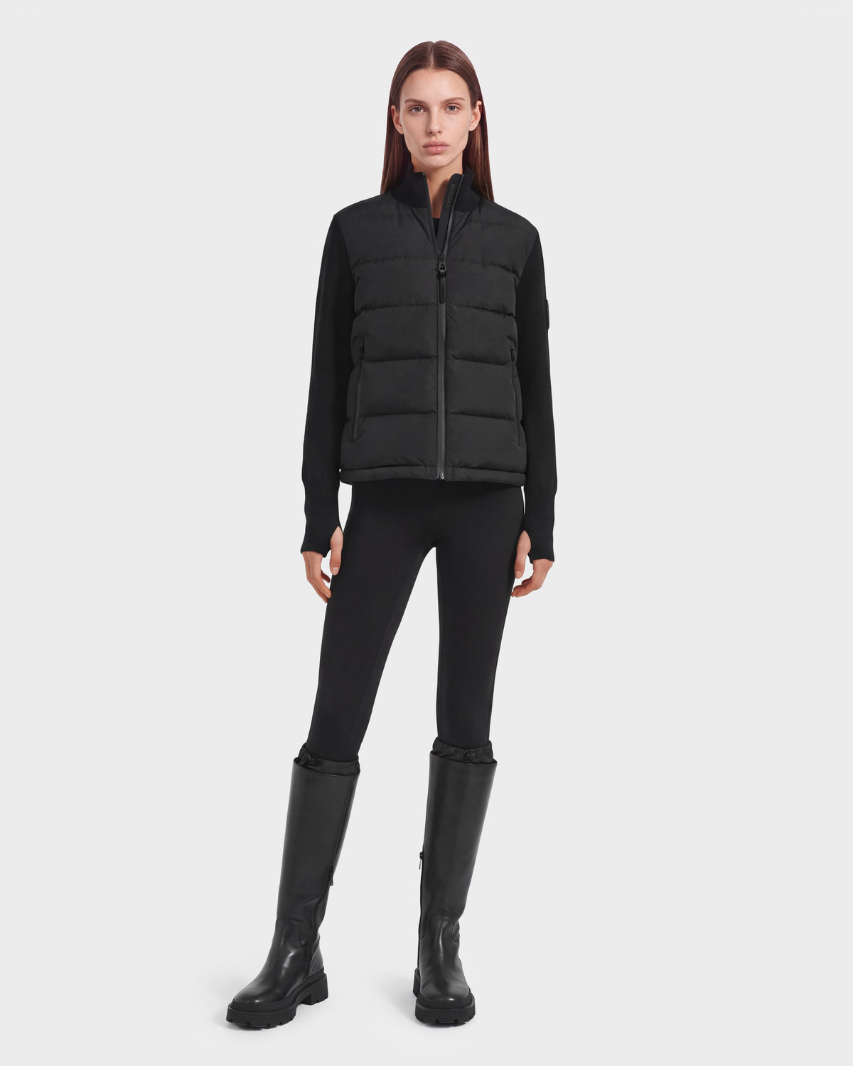 A person stands facing forward against a plain white background, wearing the limited-edition Rudsak CAMILLE HV - H125500 Black down jacket layered over a black long-sleeve top, black leggings, and tall black boots.