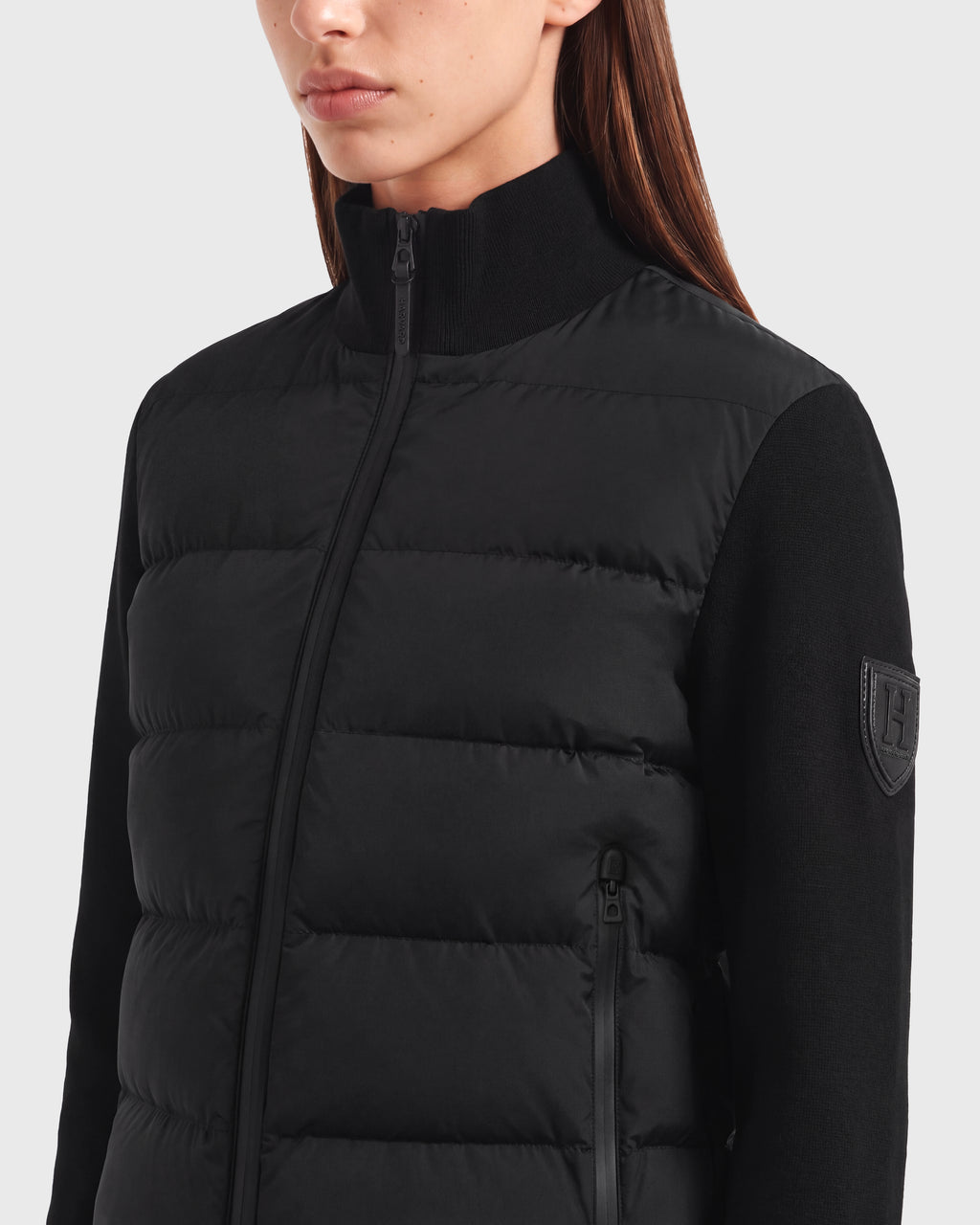 A woman wearing the Rudsak CAMILLE HV - H125500 Black jacket, a limited-edition down piece featuring a high collar, visible zipper, and signature patch logo on the sleeve, is shown from shoulders to mid-chest against a light gray background; this exclusive jacket blends modern style with unique appeal.