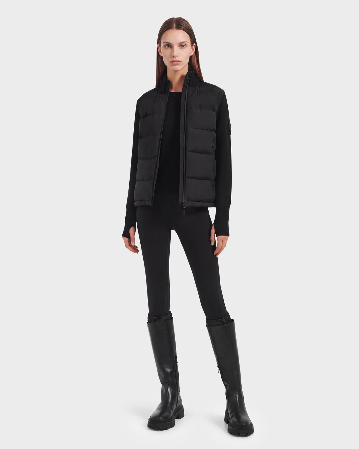 A person with straight brown hair wears the Rudsak CAMILLE HV - H125500 Black limited-edition down puffer vest, paired with a black sweater, black leggings, and knee-high black boots, standing against a plain white background.