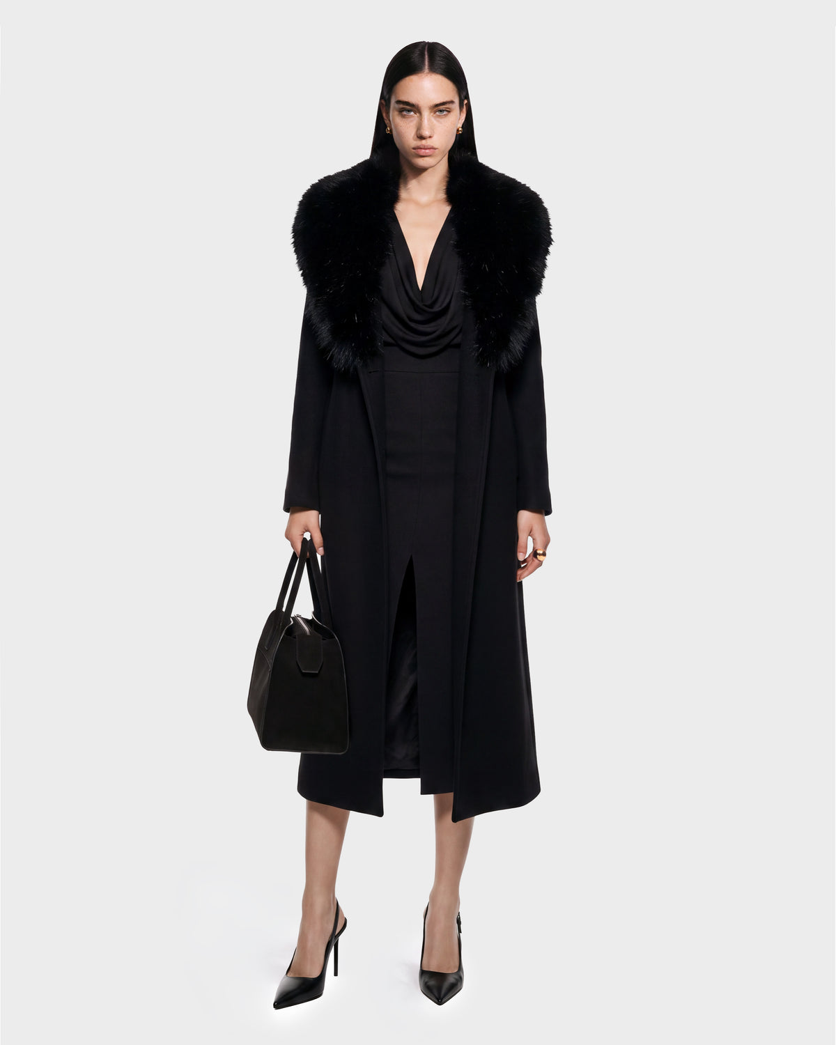 A woman stands facing forward wearing the CINDY - 8126526 Black coat by Rudsak, featuring a fur collar, paired with a black dress with a front slit, black pointed heels, and holding a black handbag. She has straight dark hair and displays a serious expression. This look is available for pre-order and is expected to ship by September 15th.